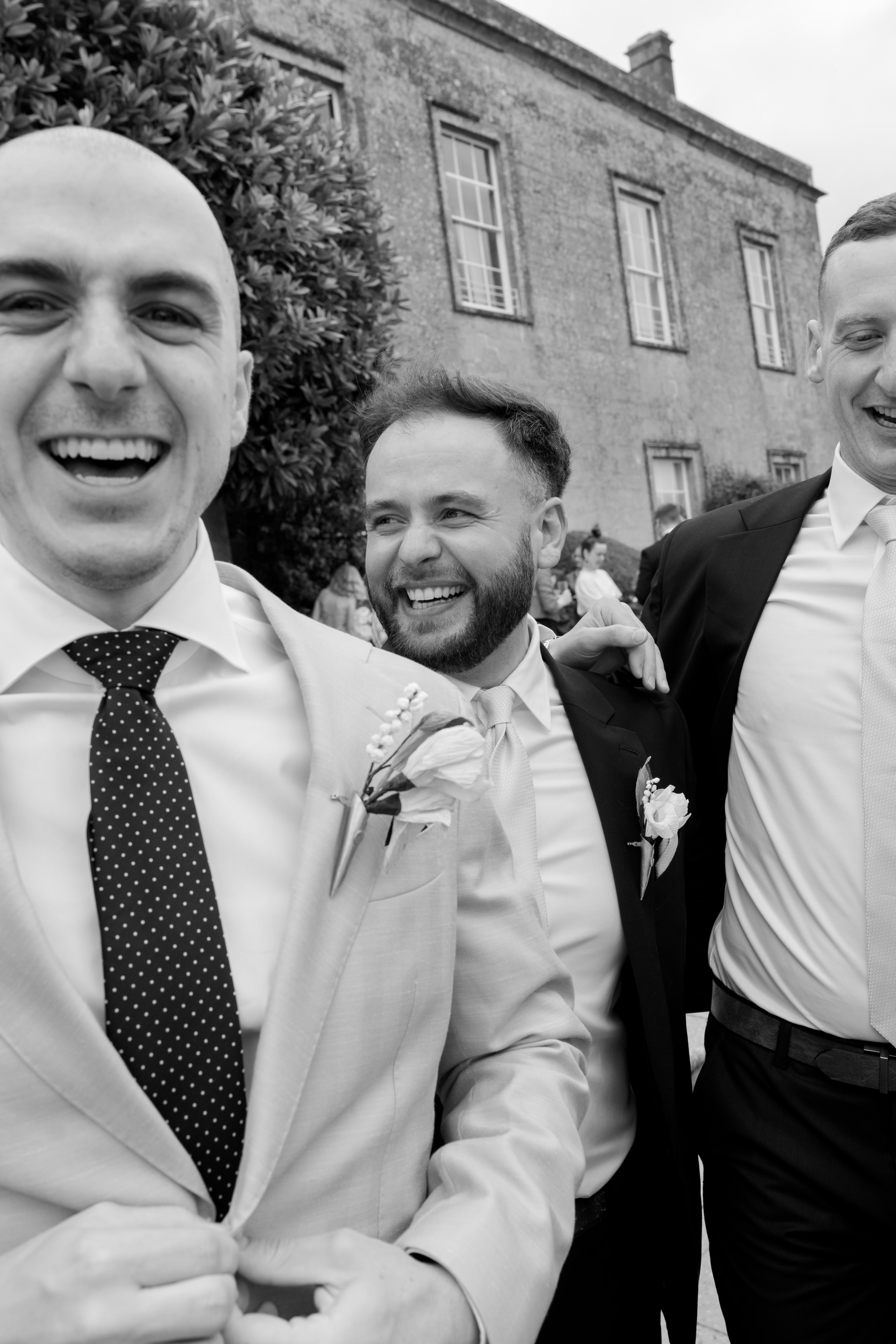 Three men wearing suits are outside, smiling and laughing with one another. There’s a large building and a leafy tree behind them.