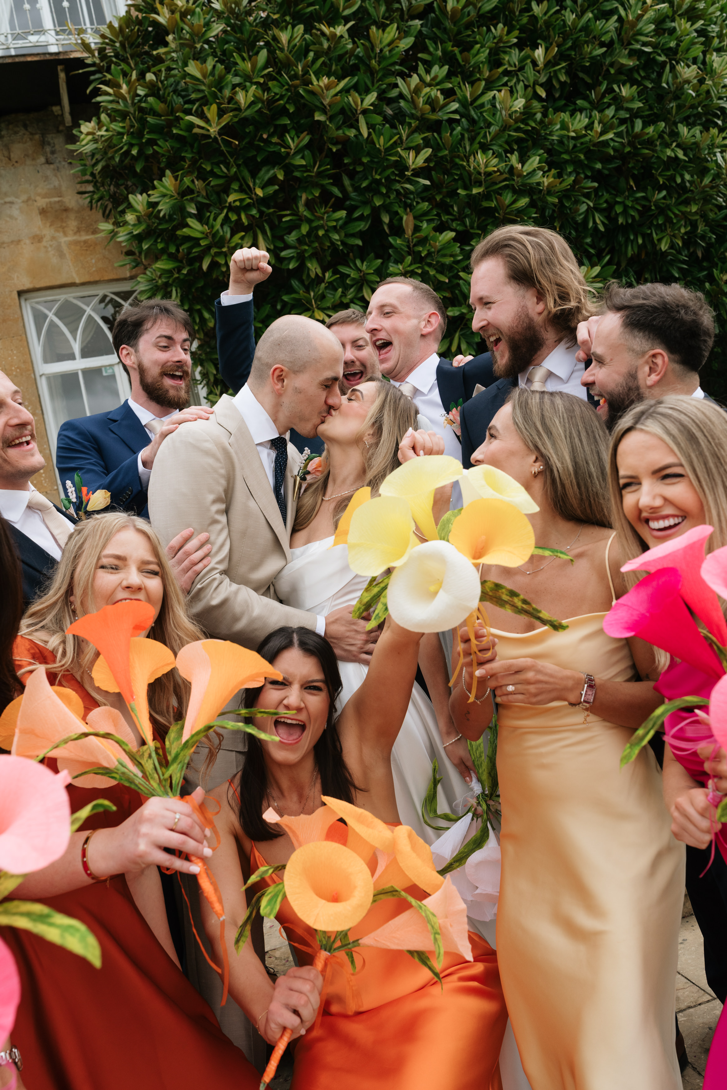 A bride and groom share a kiss outside, surrounded by happy guests who are smiling, celebrating, and holding colourful flowers.