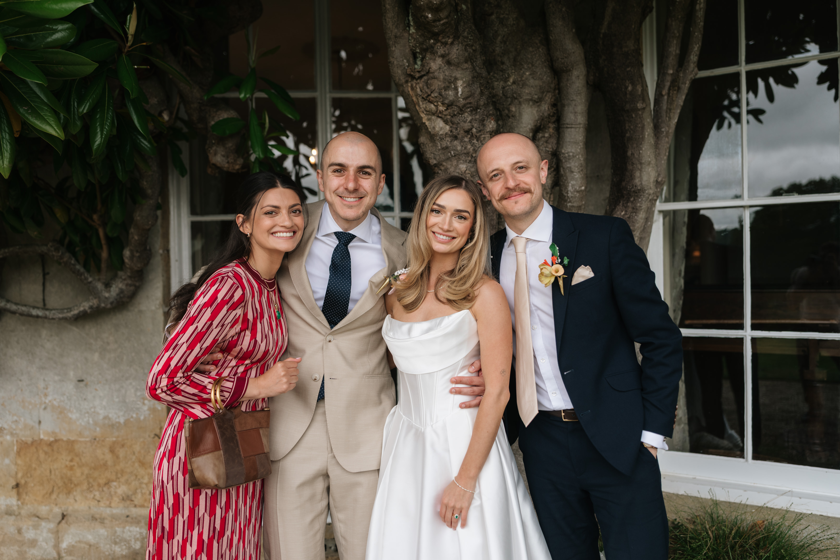Four adults are standing together and smiling in front of a window and a tree. They’re all dressed up: one woman is wearing a white dress, two men are wearing suits, and another woman is wearing a red patterned dress.