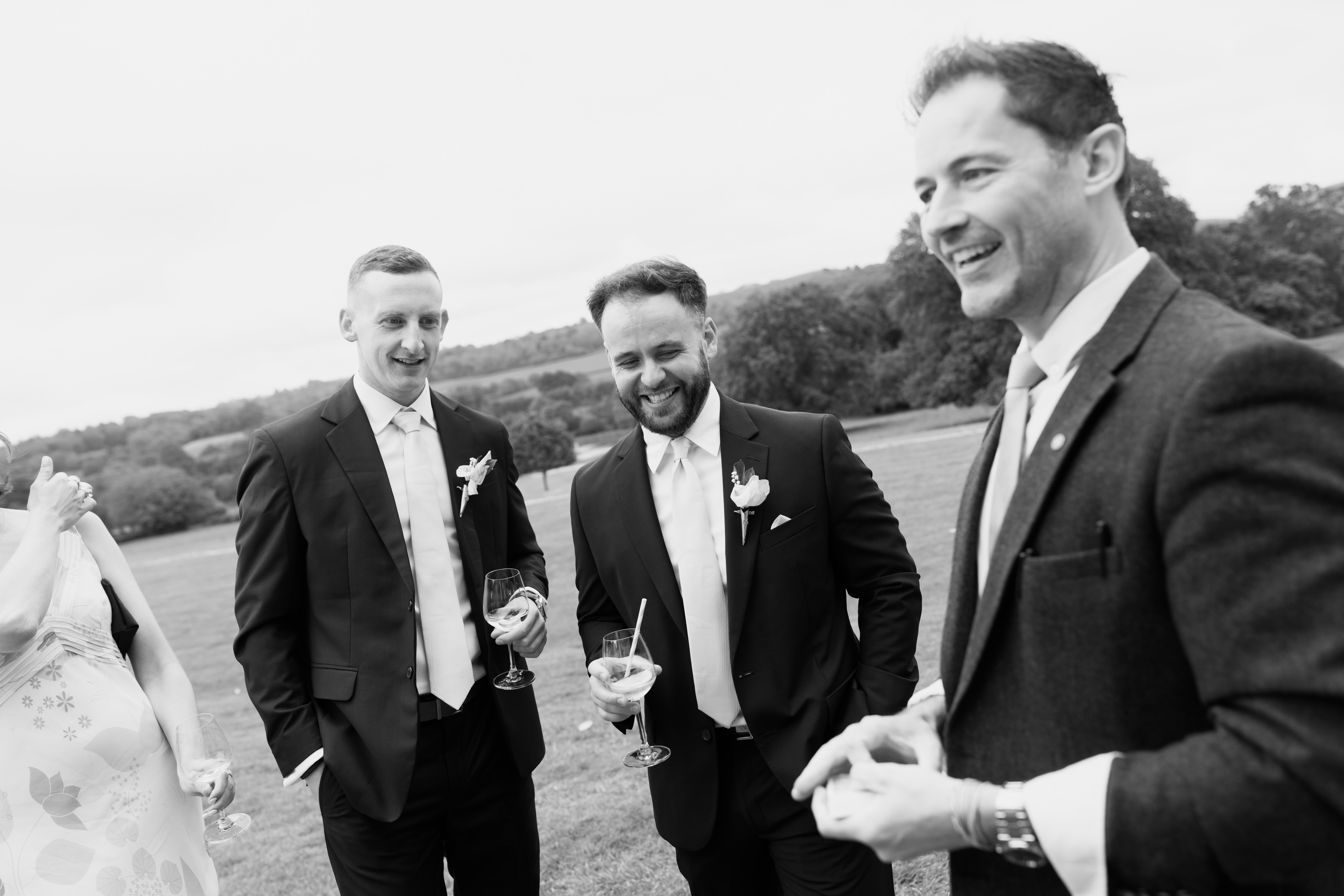 Three men wearing suits are standing on the grass outside, holding drinks and smiling as they talk with other people at a posh event.