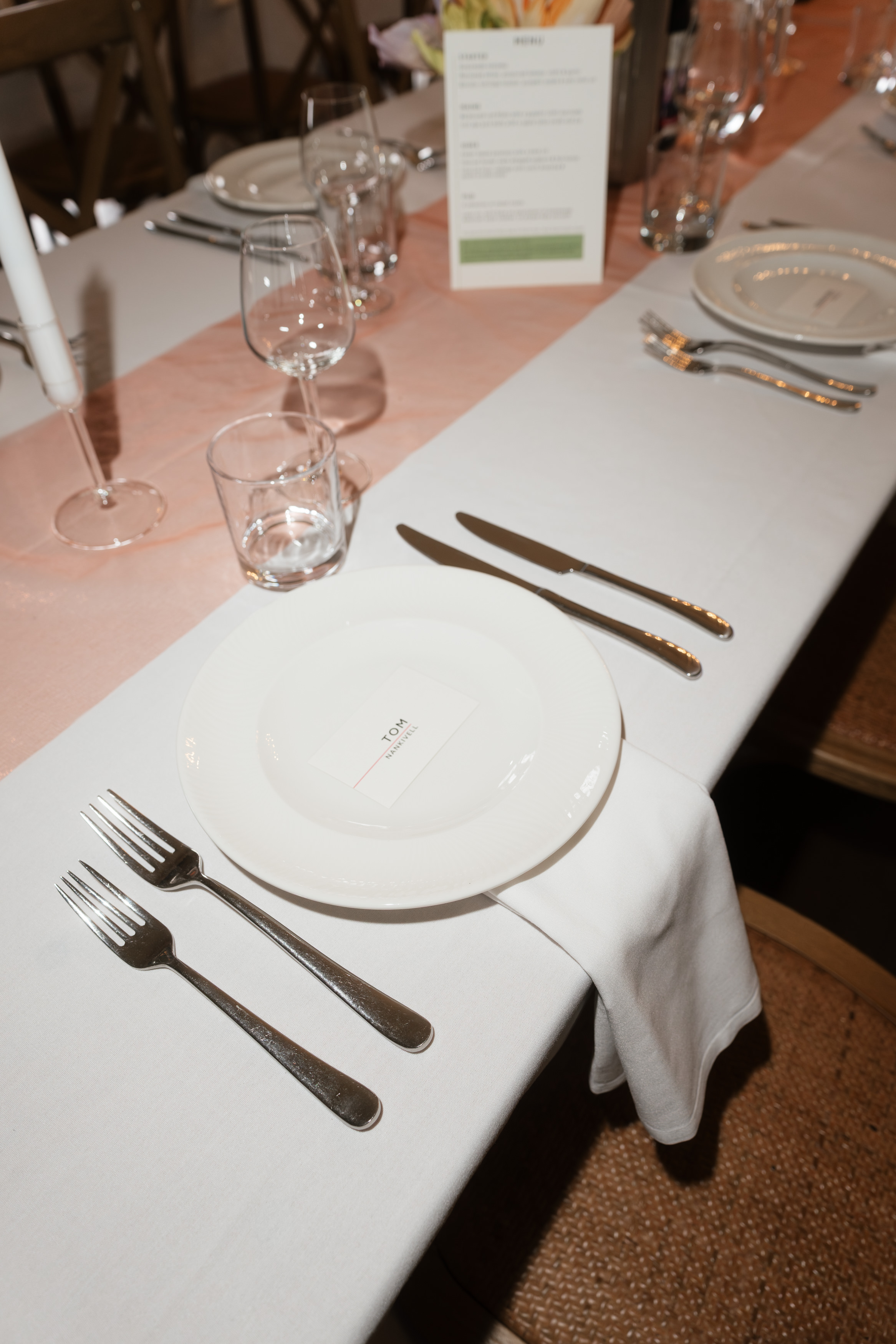 A table laid for a meal with a white plate, cutlery, a wine glass, and a menu card on top of a white tablecloth and a peach-coloured runner.