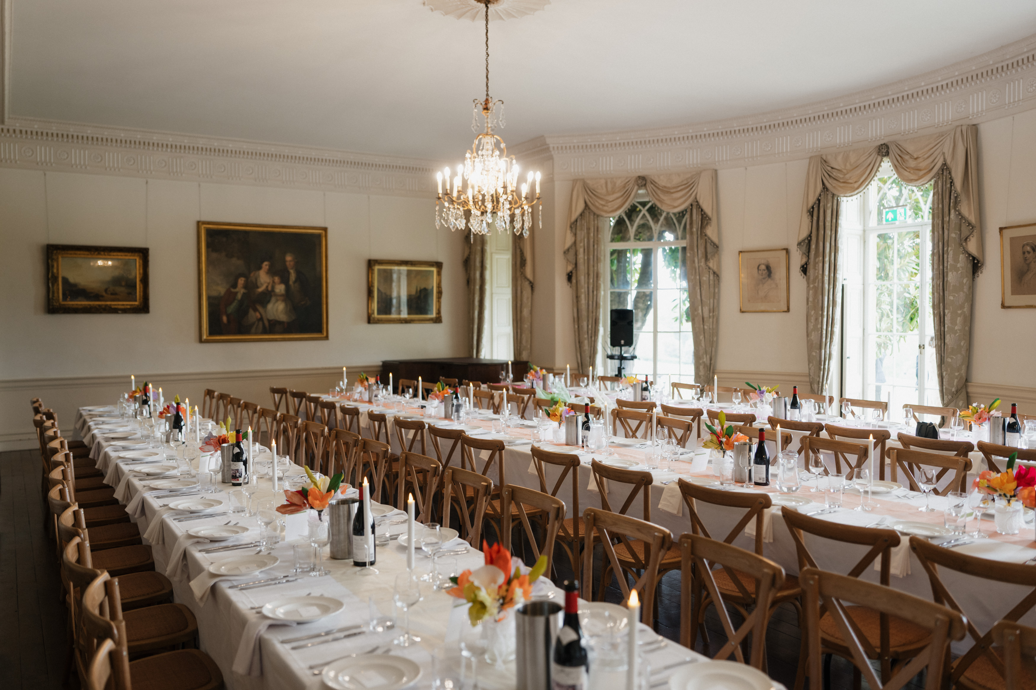A fancy dining room with two long tables ready for a gathering, covered in white tablecloths and set with wine bottles, glasses, and flowers. Chandeliers hang from the ceiling and paintings are on the walls, with plenty of natural light filling the elegant room.