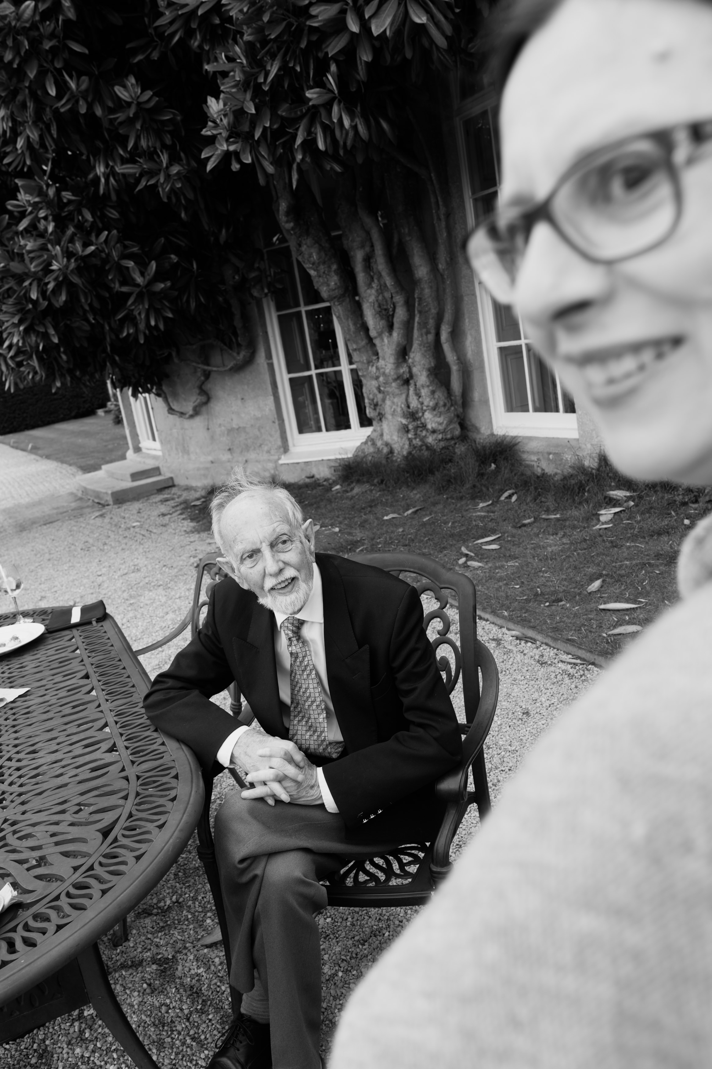 An older man is talking at an outdoor table while a woman wearing glasses smiles in front.