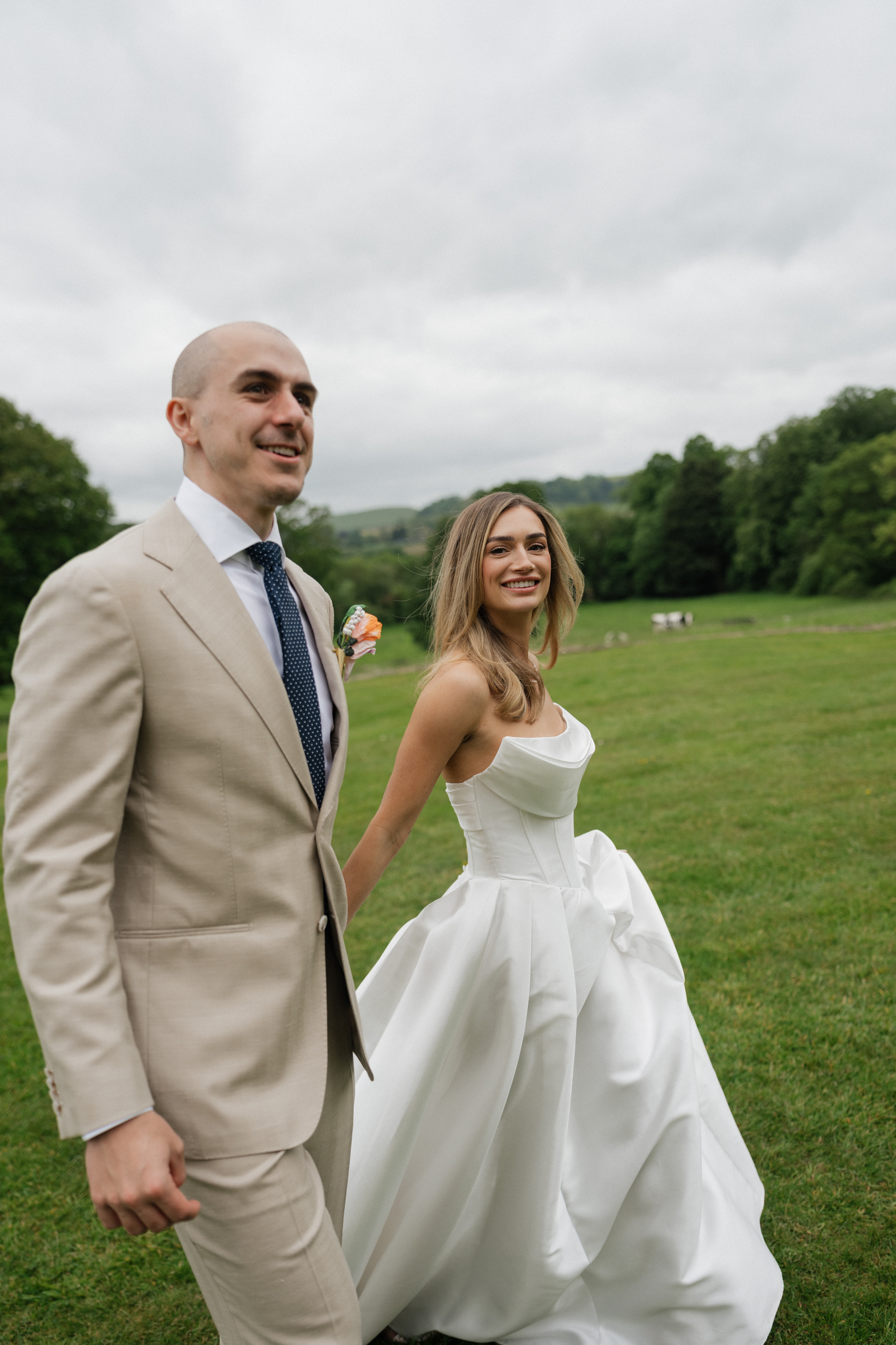 A bride wearing a white dress and a groom in a beige suit are walking together, holding hands on a grassy field with trees behind them.
