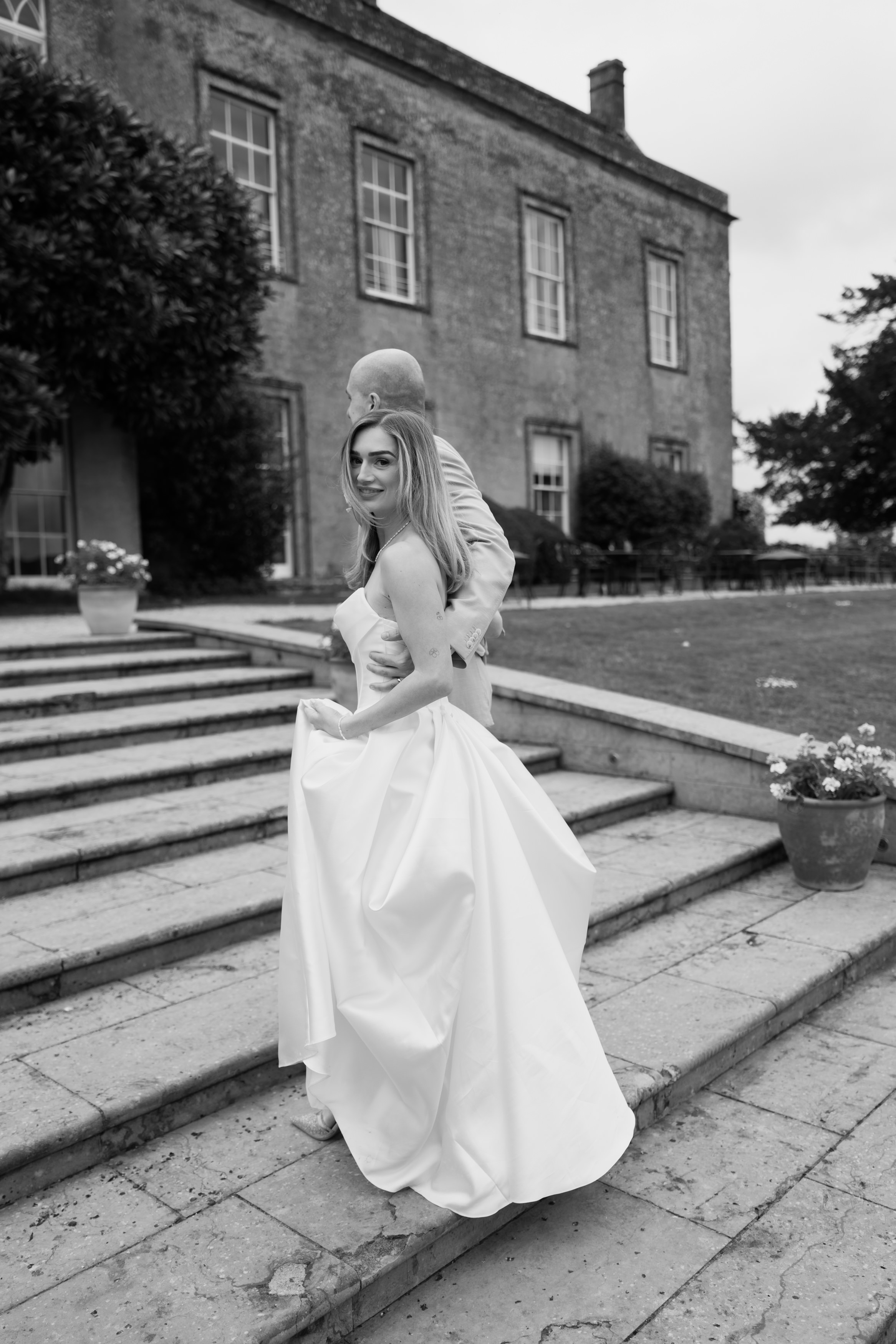 A woman wearing a wedding dress stands on some stone steps outside, glancing back over her shoulder. There’s a man in a suit next to her, and a big building behind them.