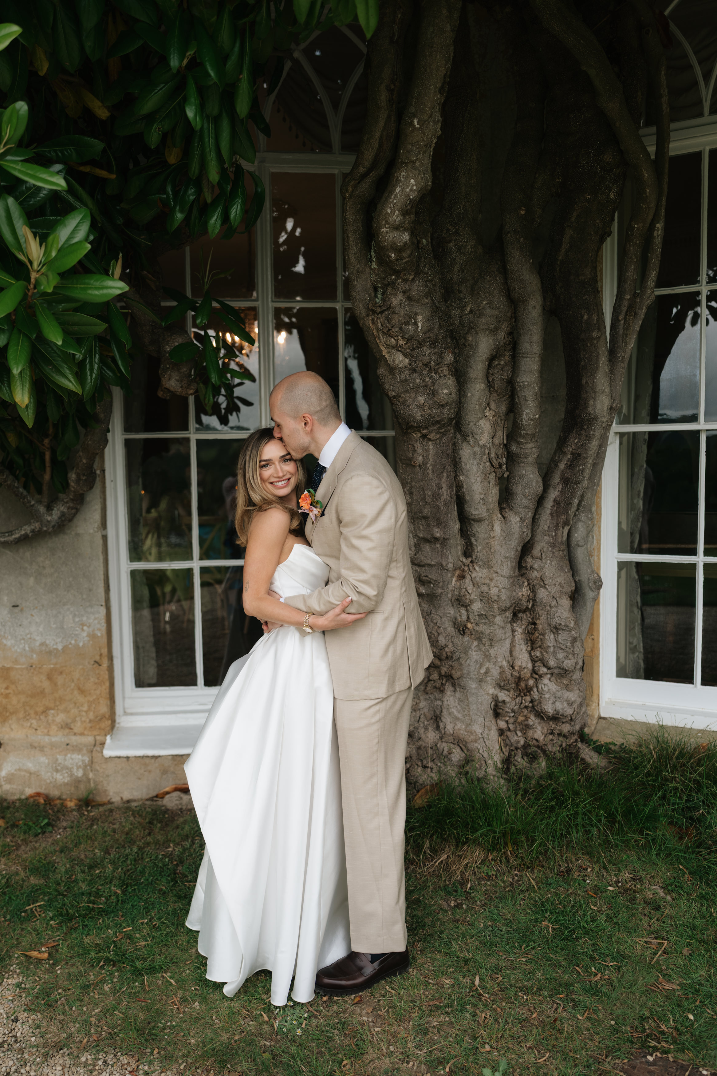 A bride and groom dressed for their wedding stand in front of a large tree and some windows, while the groom kisses the bride on her forehead.