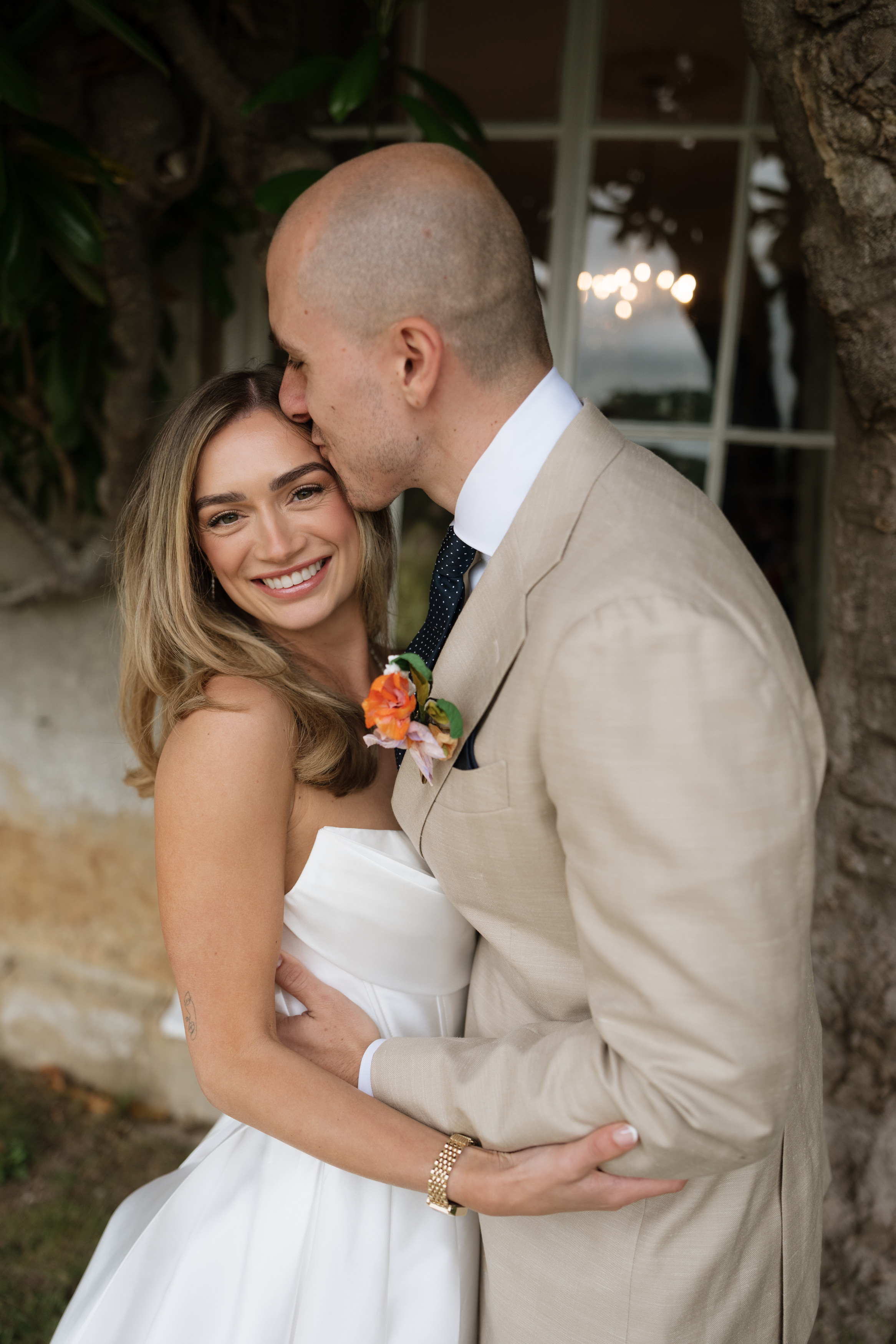 A man wearing a beige suit gives a forehead kiss to a smiling woman in a white dress whilst they hug outside next to a tree.