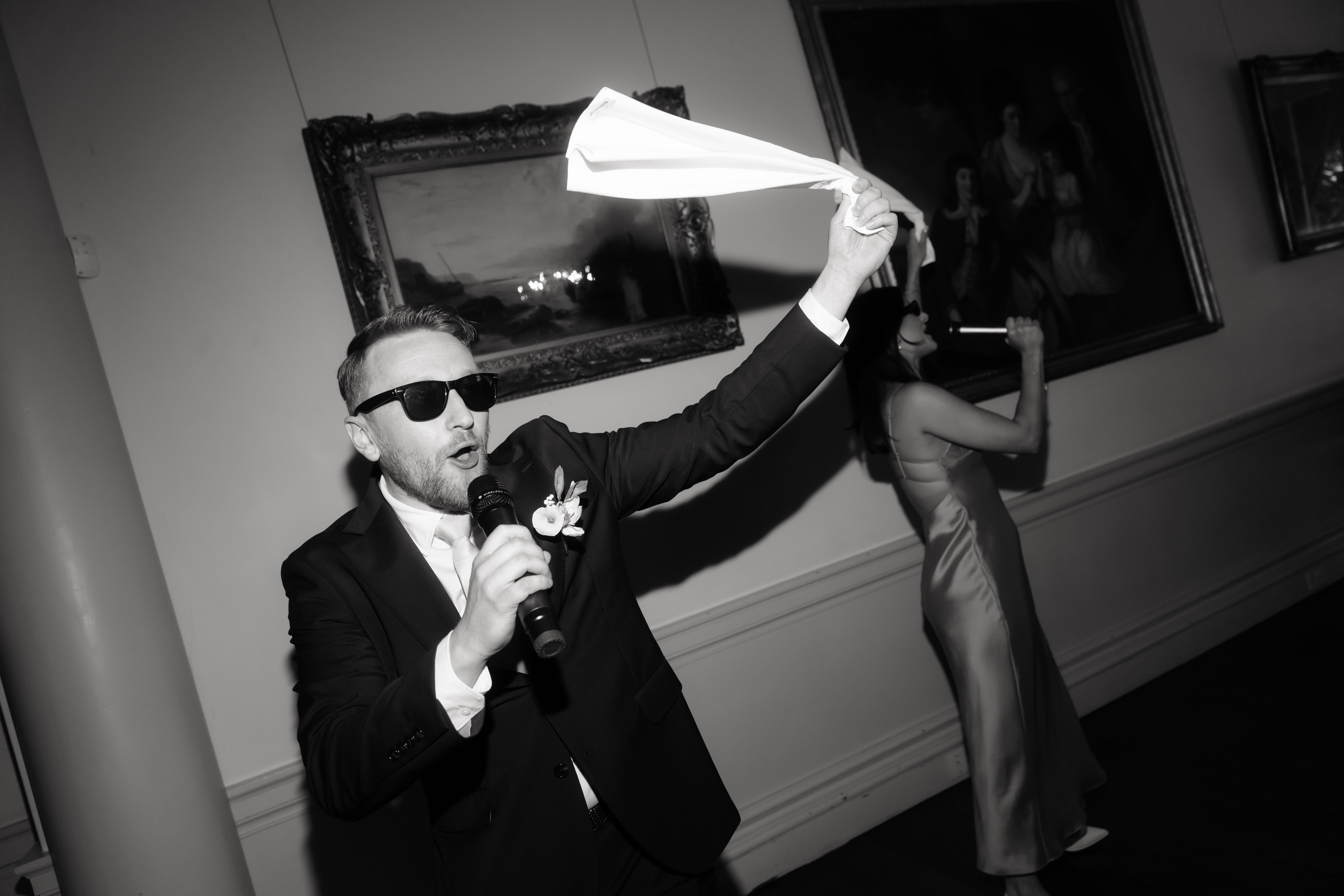 A man wearing a suit and sunglasses is holding a microphone and waving a serviette, while a woman in a dress stands behind him in a room decorated with framed paintings.