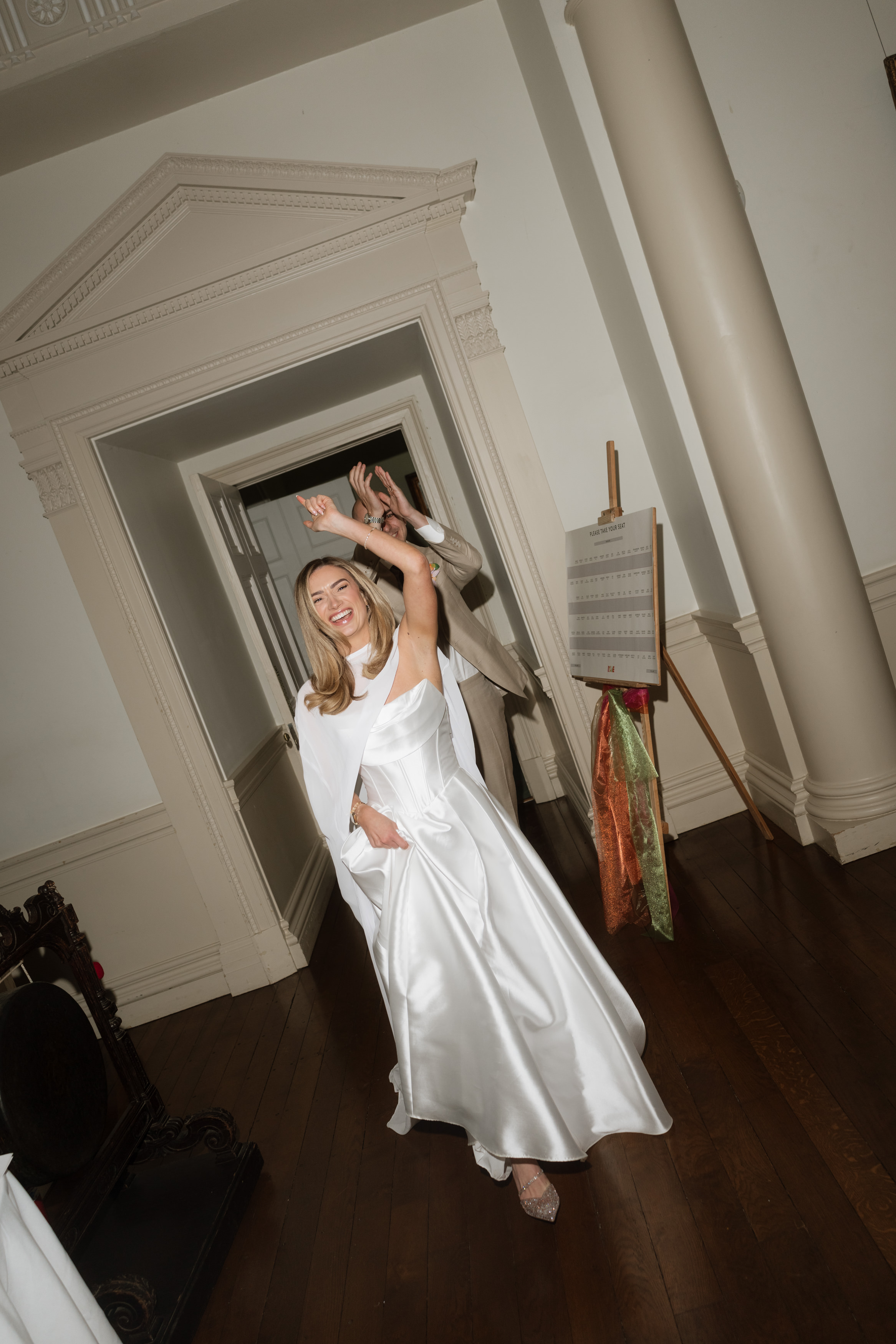 A woman wearing a white dress is smiling and dancing in a corridor with wooden floors and white walls, holding her arms up.