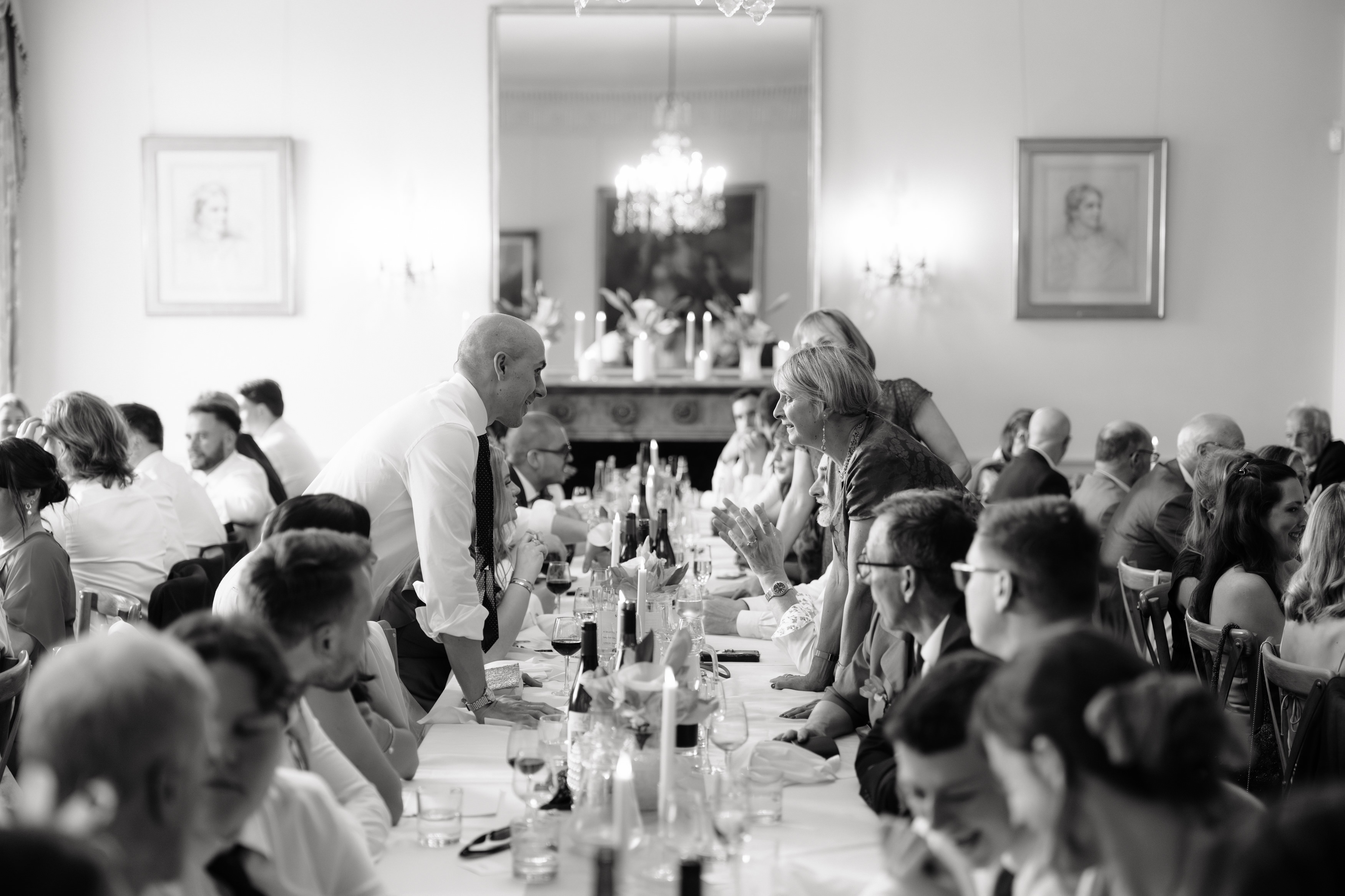 A black and white photo shows a large group of people sitting at a long table, with two people standing in the middle and talking to each other across the table.