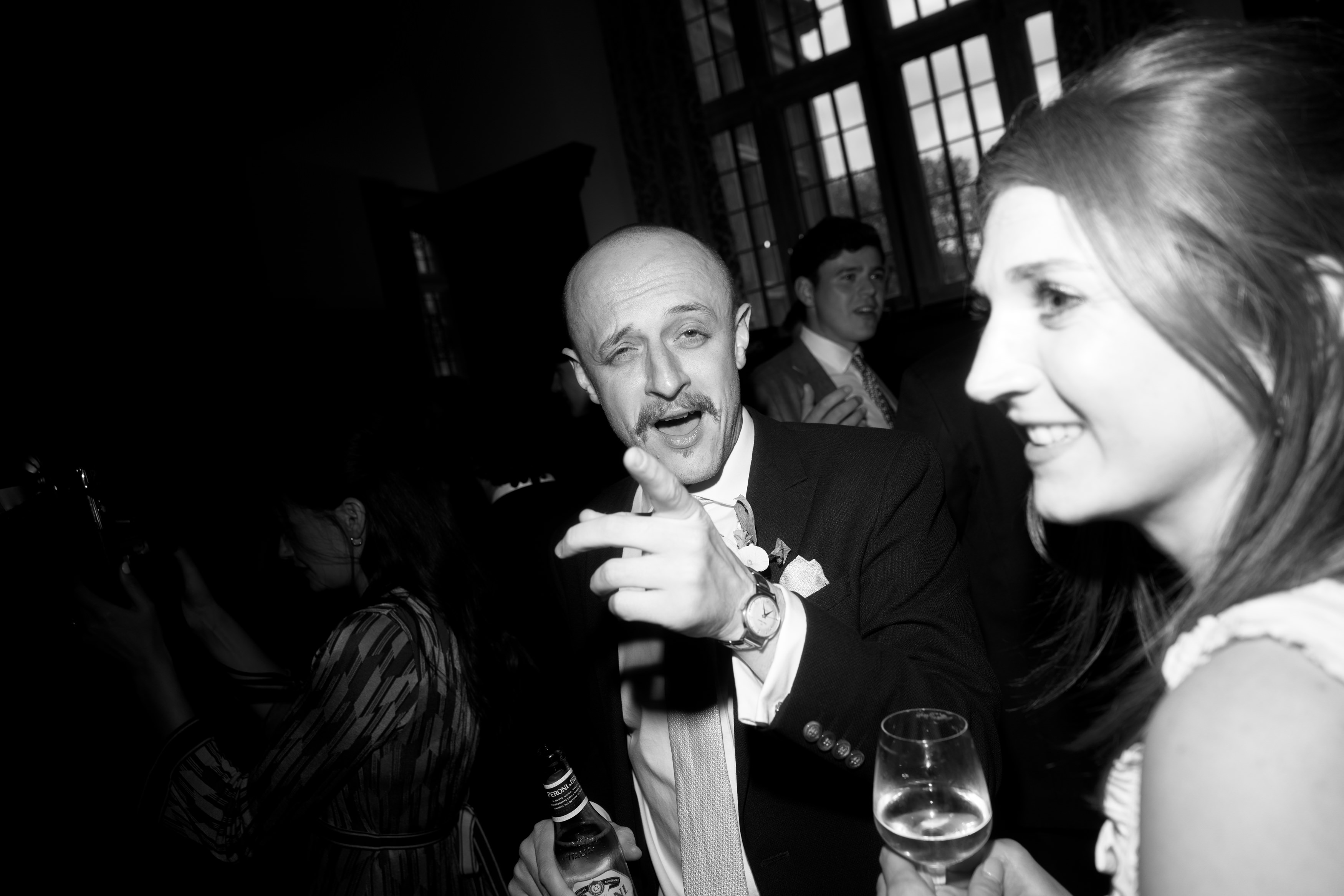 A man in a suit is holding a drink and gesturing whilst standing next to a smiling woman who has a glass of wine, both at a social event.