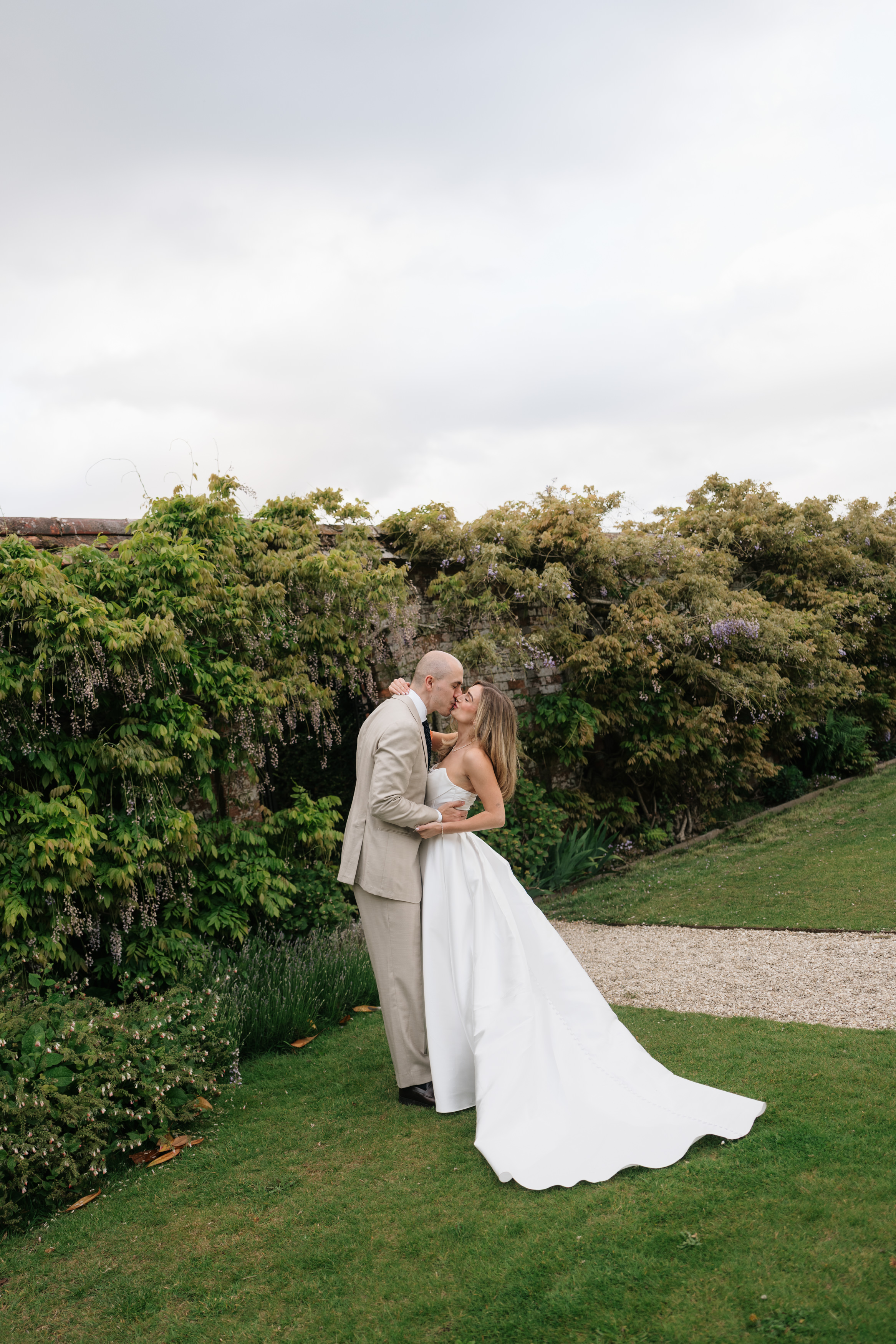 A bride and groom are hugging and kissing on a grassy lawn, with plants and flowers behind them and a cloudy sky overhead.