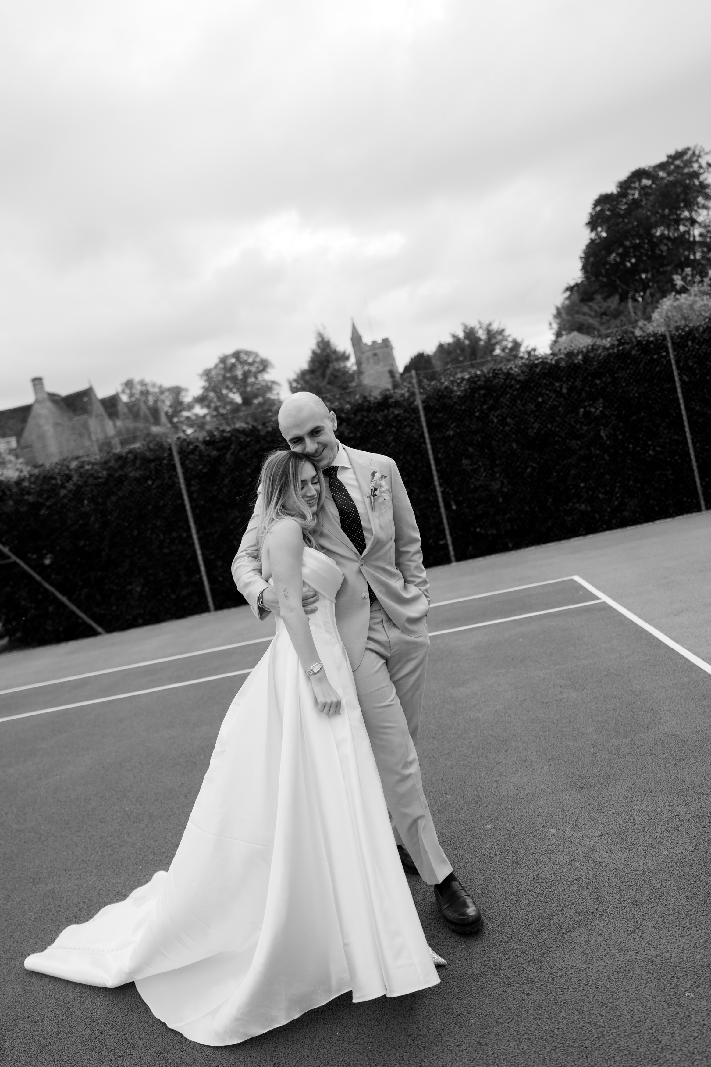 A couple in wedding clothes stand together on an outdoor tennis court, with trees and some buildings behind them.