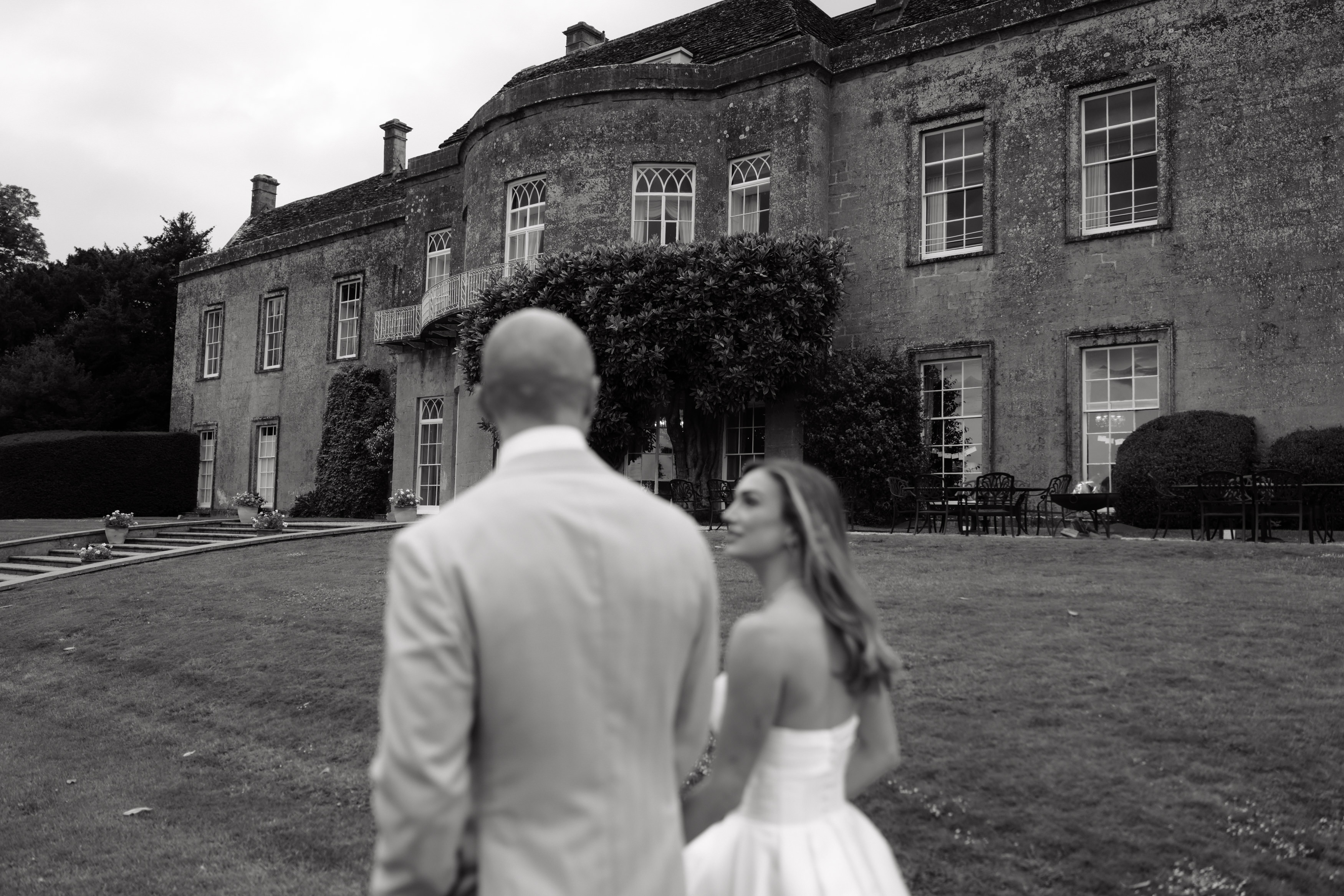 A couple dressed up in formal clothes walks across the grass towards a big old building, shown from behind.