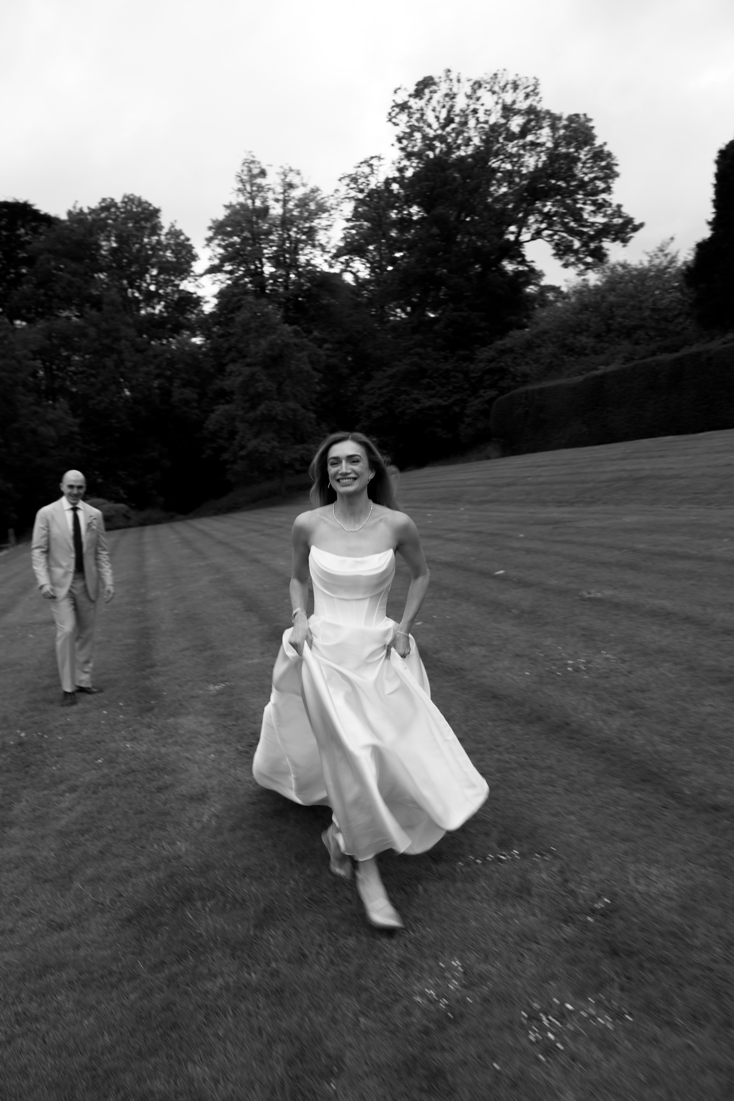 A woman wearing a white dress is running on the grass and smiling, with a man in a suit walking behind her. There are trees and hedges in the background.