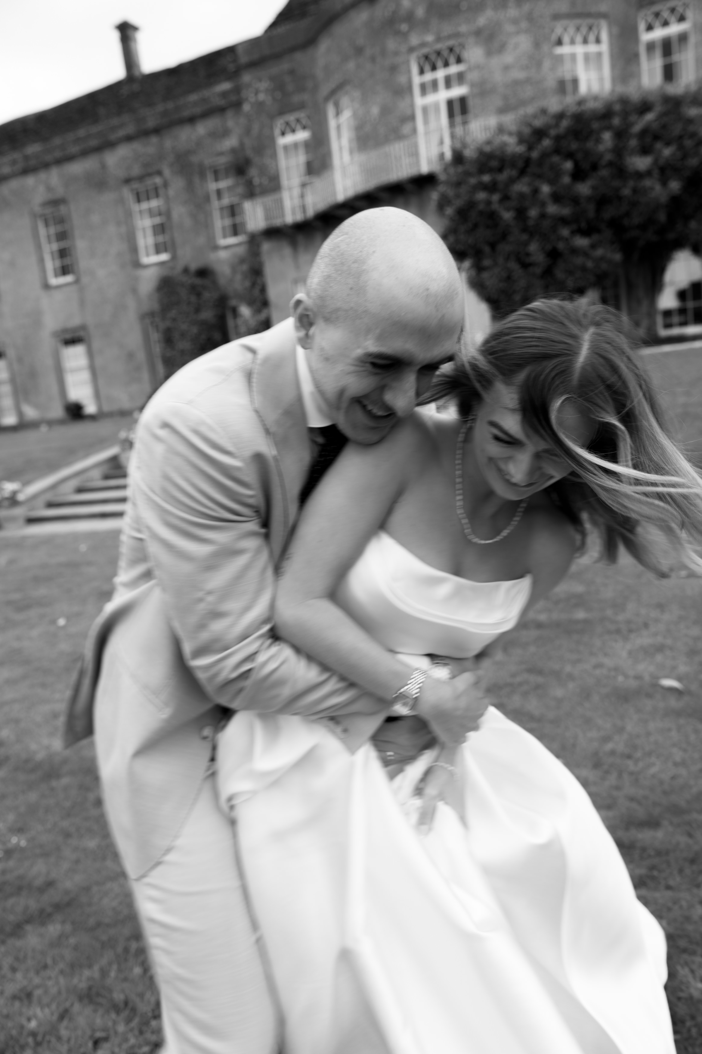 A man wearing a light-coloured suit is hugging a woman in a strapless dress outside near a big building. They’re both smiling, and the black-and-white photo looks a little blurry, as if they were moving.