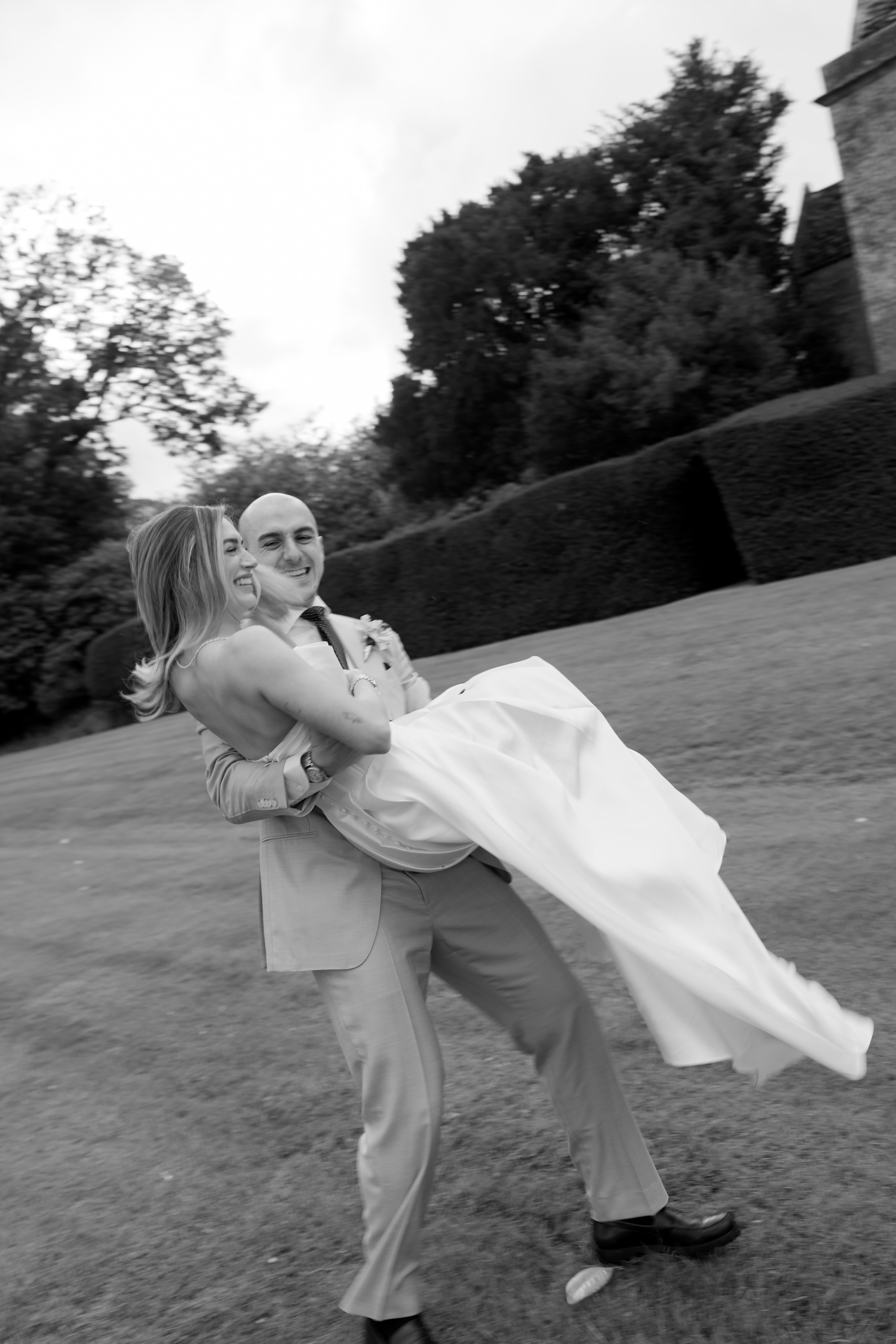 A man wearing a suit is carrying a woman in a wedding dress across the grass, and they’re both smiling. The picture is in black and white, and there are trees and bushes behind them.