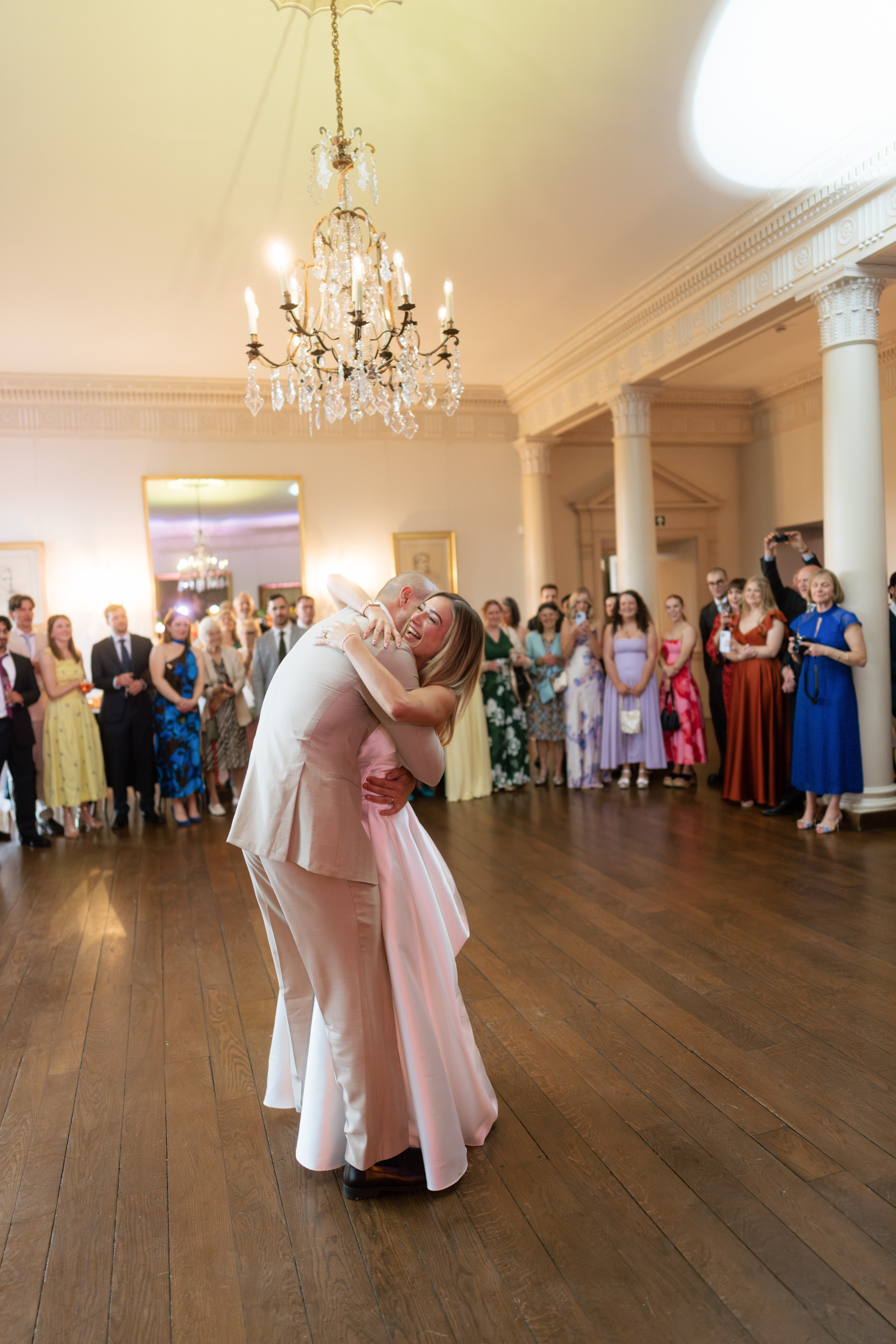 A couple dressed up in smart clothes hugs and dances in the middle of a ballroom, while people around them watch and take photographs.