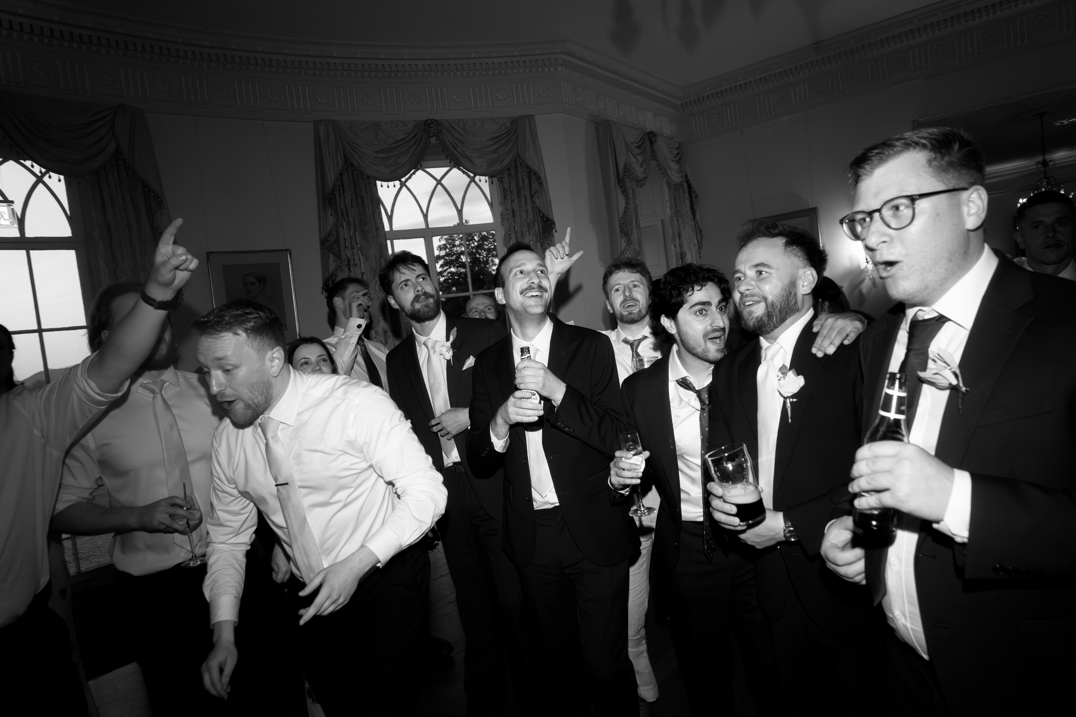 A group of men in suits are indoors, singing and celebrating, holding drinks and looking excited in what seems like a posh place.