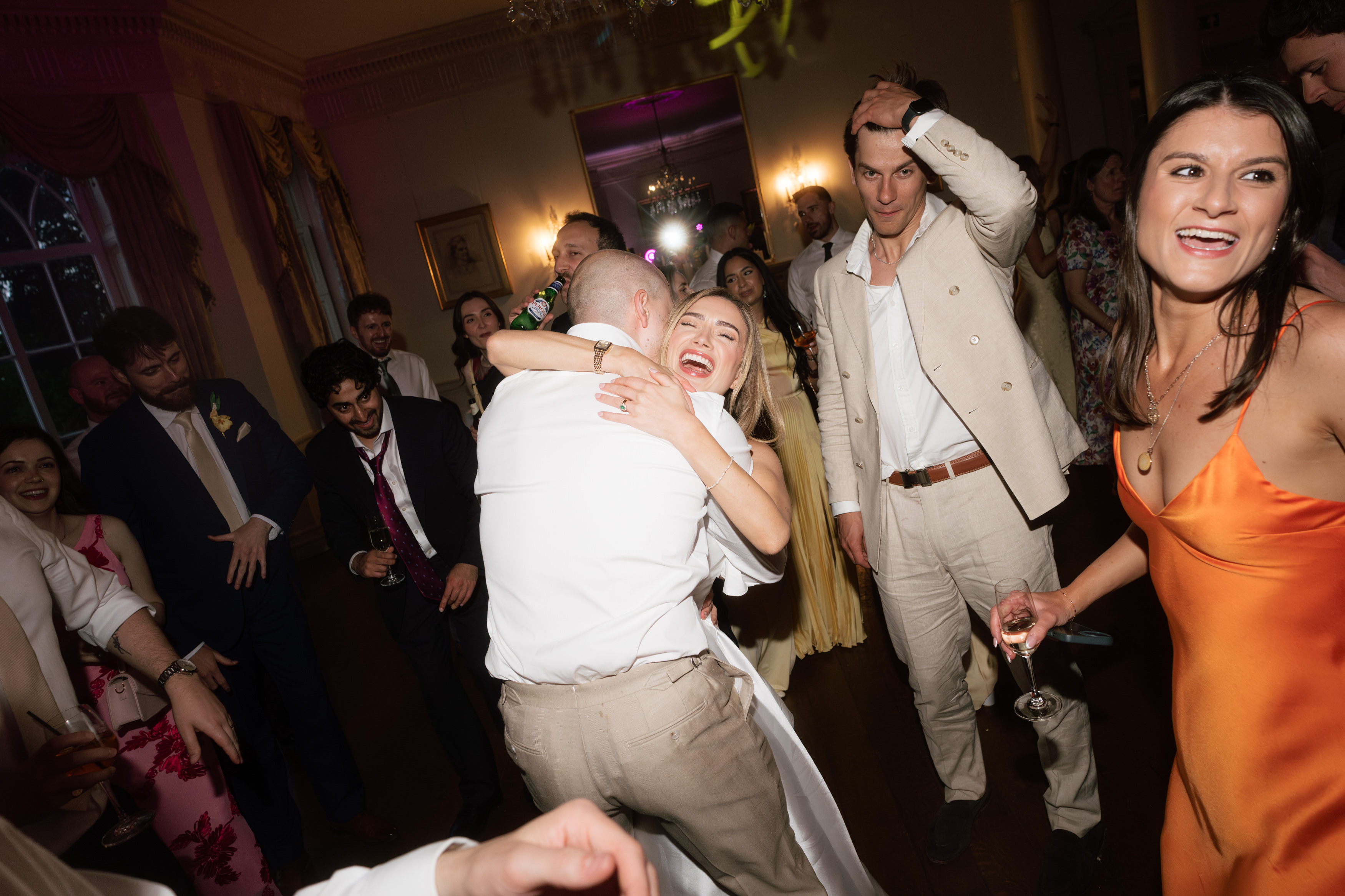 People dressed up nicely are dancing and having fun at an indoor party. One couple is hugging, and others are smiling and holding drinks.