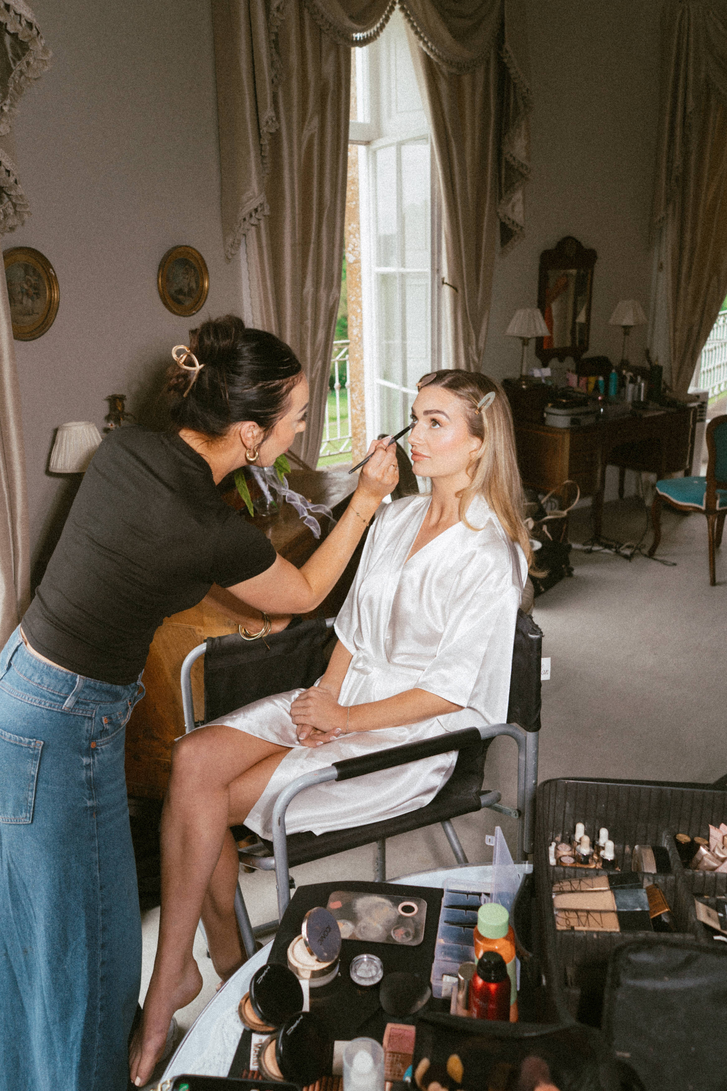 A woman wearing a white dressing gown is sitting in a chair whilst someone does her make-up in a bright room, with beauty products on a nearby table.