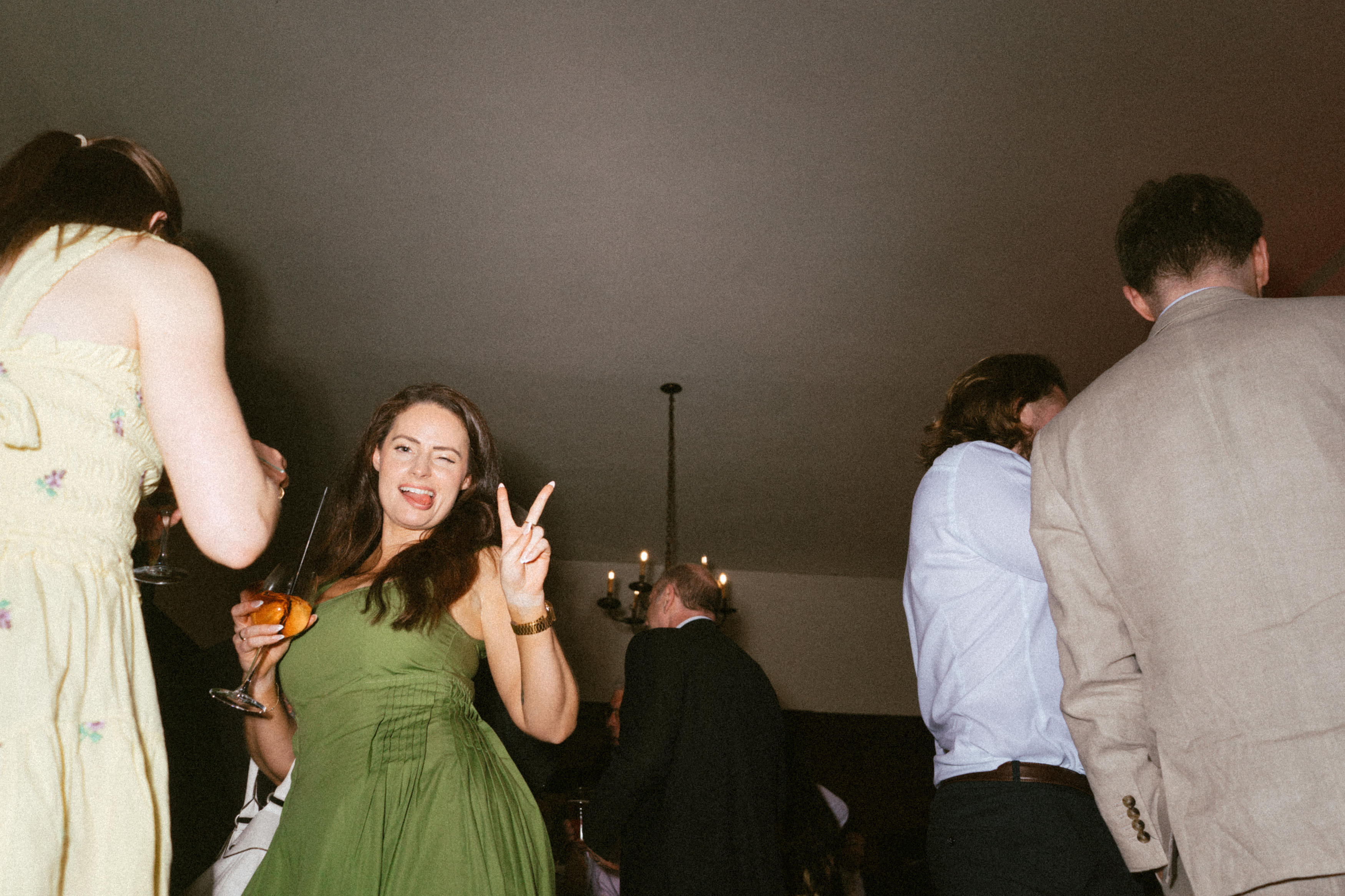A woman wearing a green dress is holding a drink and flashing a peace sign at a party, with other people dressed up and standing around her.