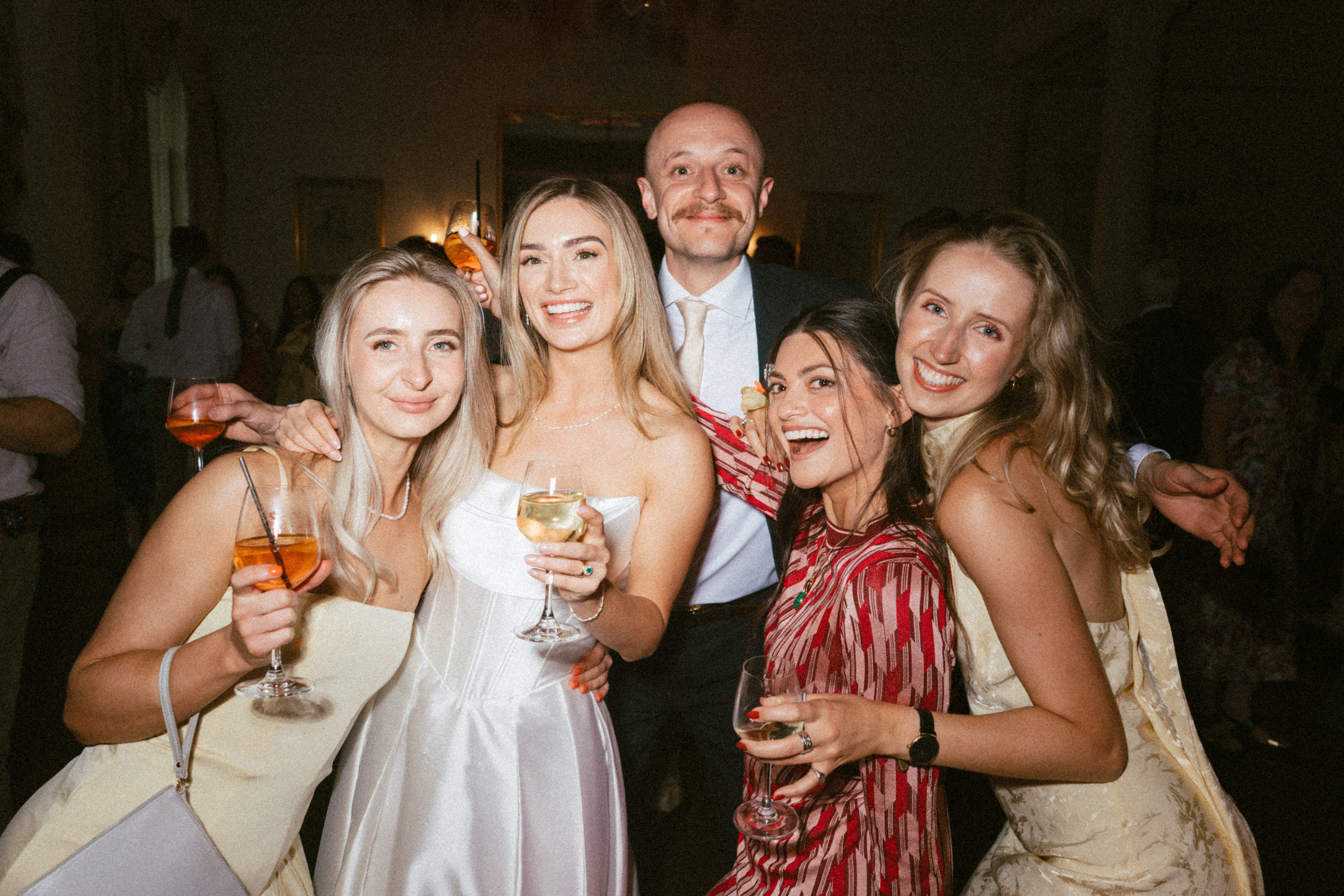 Five adults wearing smart clothes are smiling and posing together at an indoor event, each holding a drink.