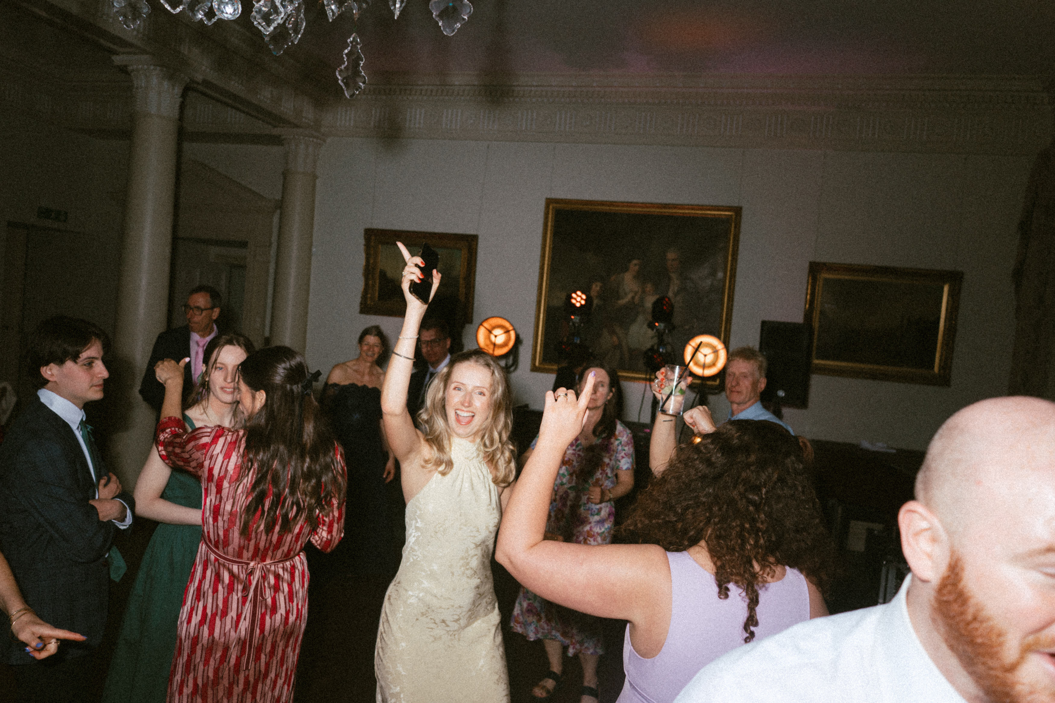 People are having fun and dancing at an indoor party, smiling with paintings on the walls and lights in the background.