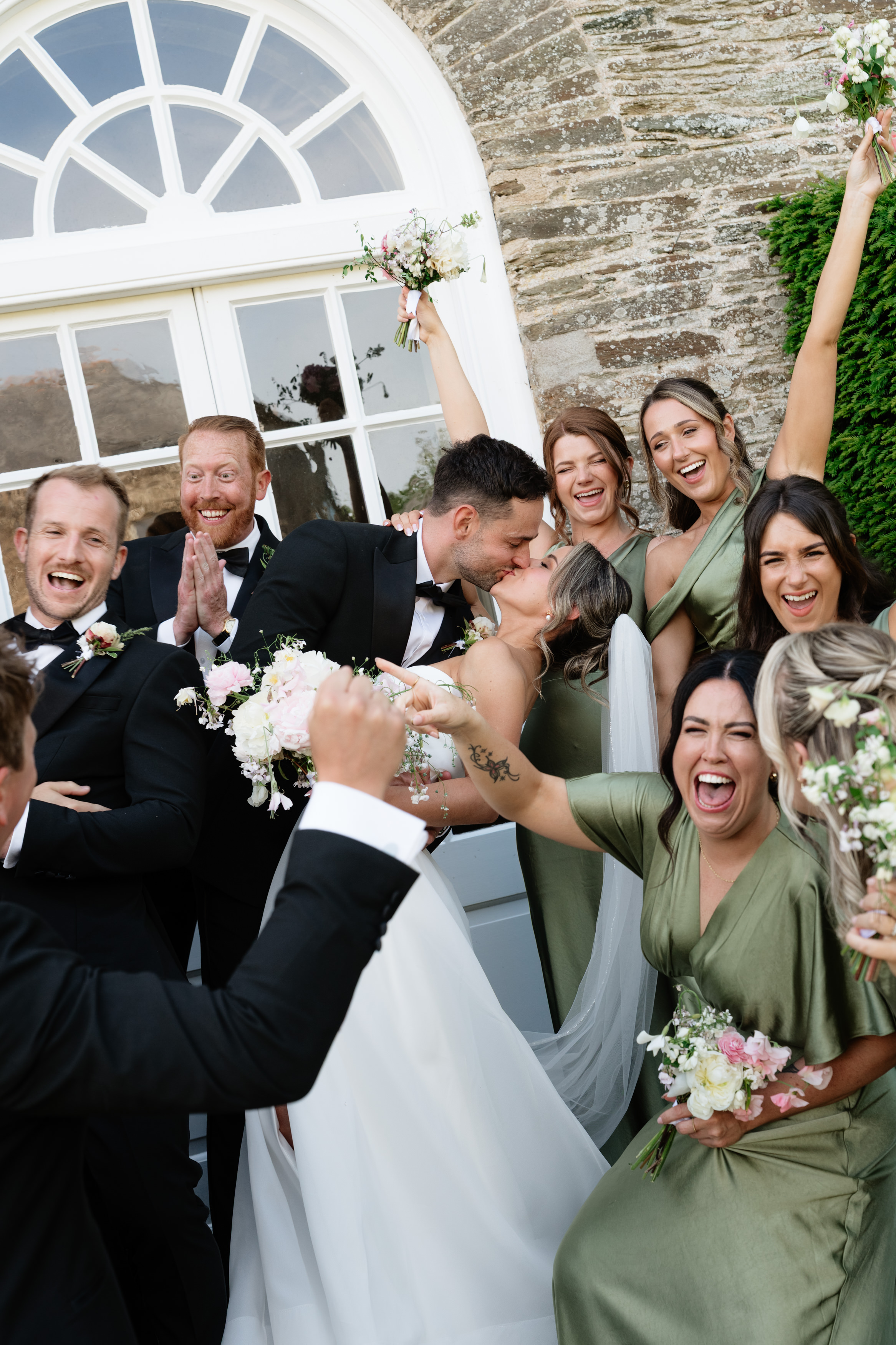 A bride and groom share a kiss whilst their wedding party, wearing green and black outfits, celebrates around them outside by a large window and stone wall. Everyone is smiling and holding bouquets.