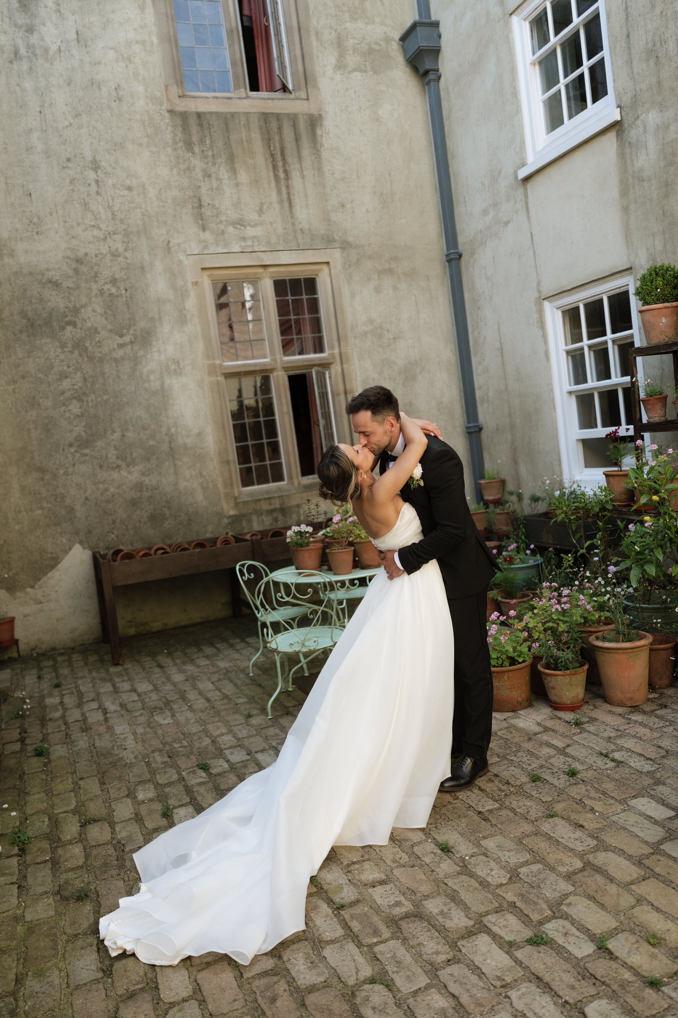 A bride and groom share a kiss in a courtyard that has potted plants and a metal bench, with old building walls and windows around them.
