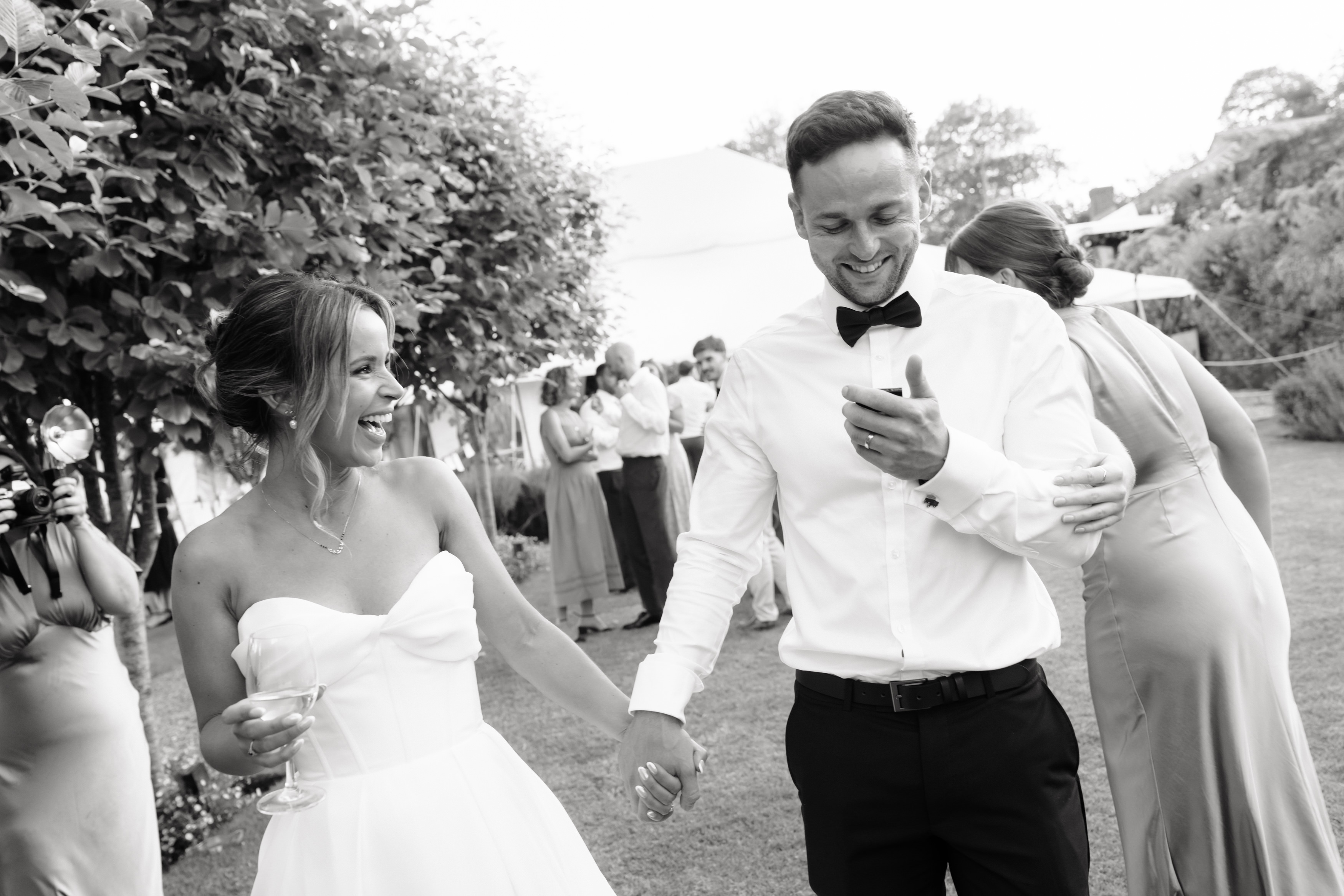The bride and groom are smiling and holding hands as they walk outside at their wedding, with guests chatting in the background.