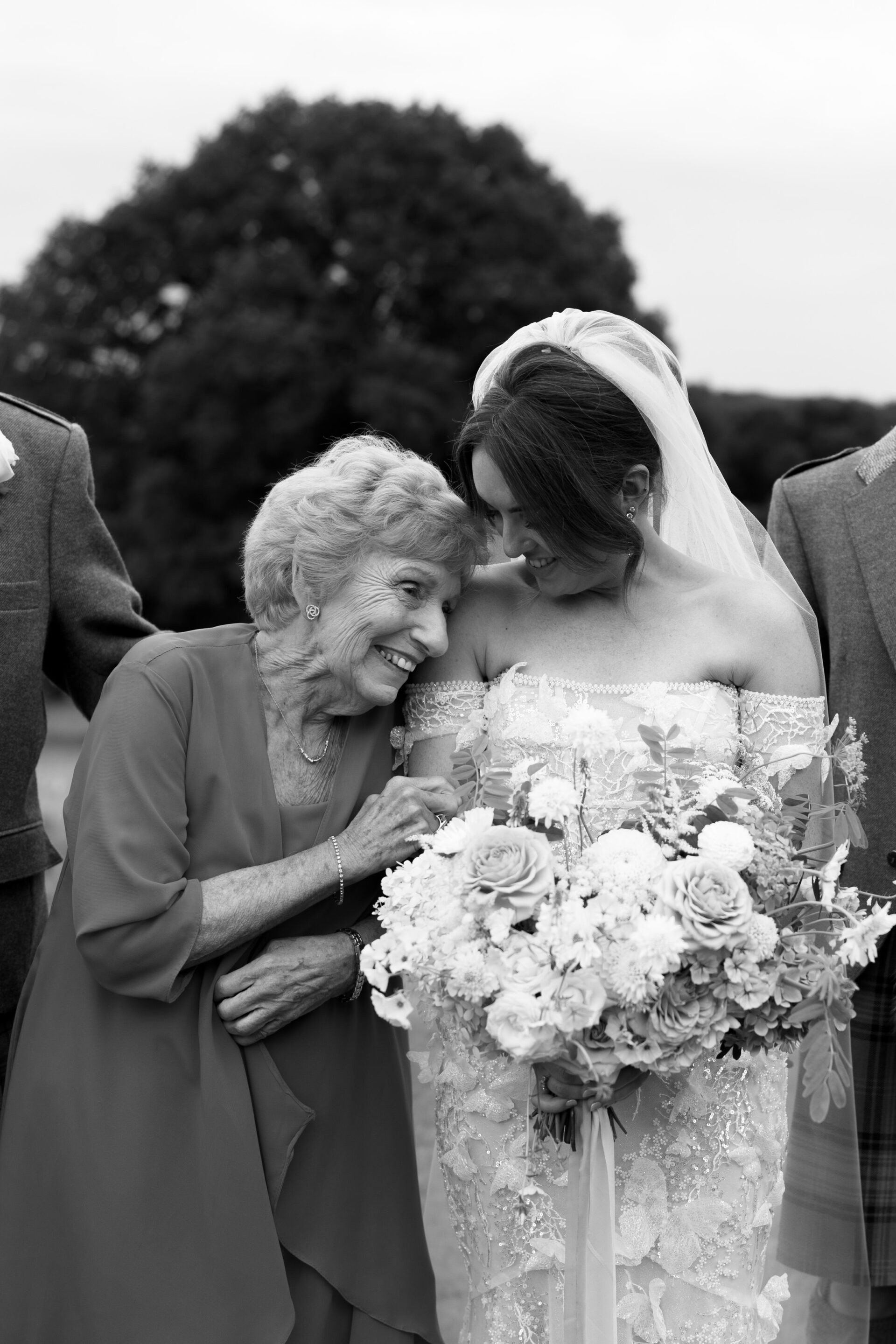 A bride wearing a strapless dress is holding a bunch of flowers. An older woman in a dress is leaning on her shoulder, smiling. They are standing outside with a tree in the background.
