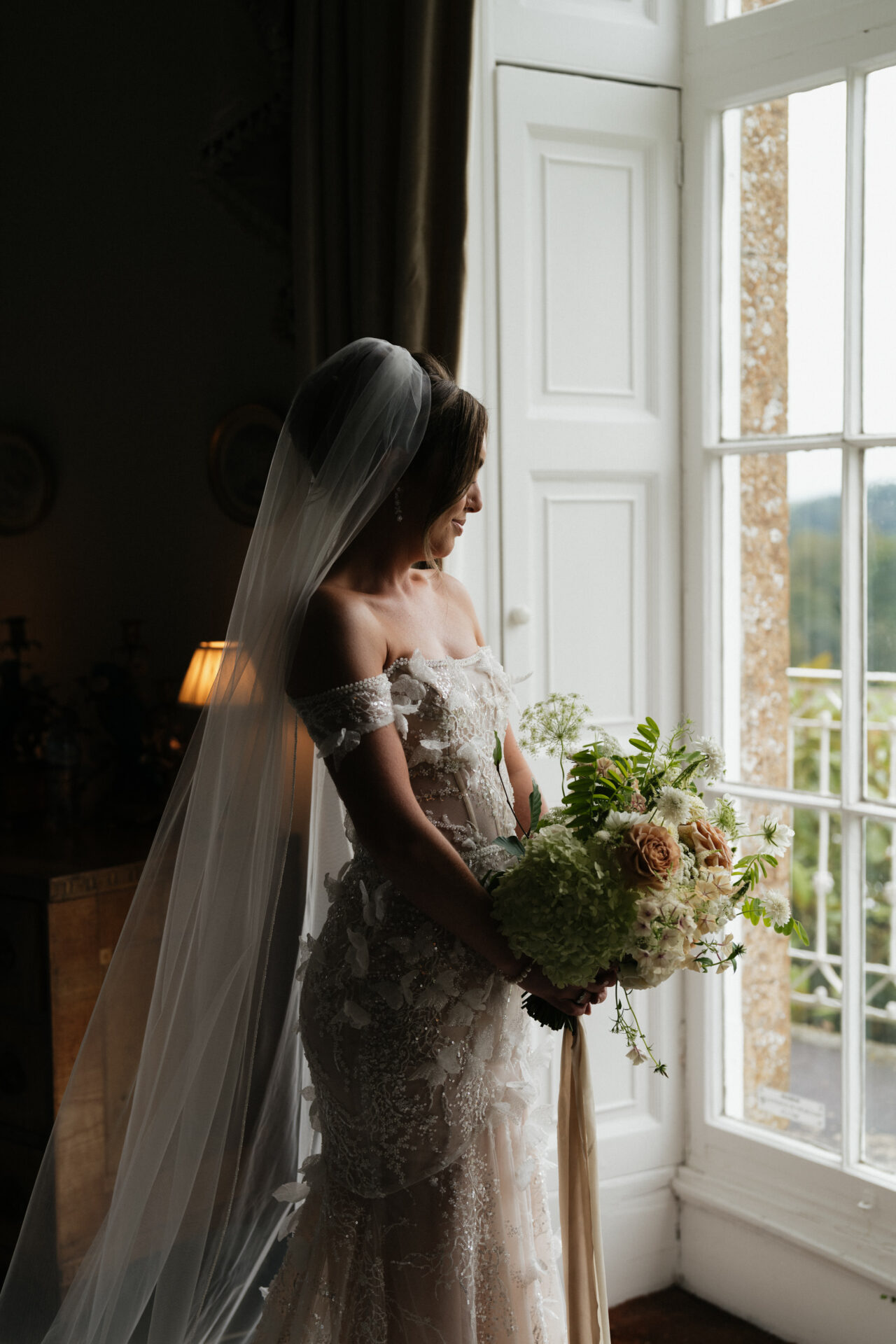 A bride in a beautiful dress and veil holds a bunch of flowers while gazing out of a large window.