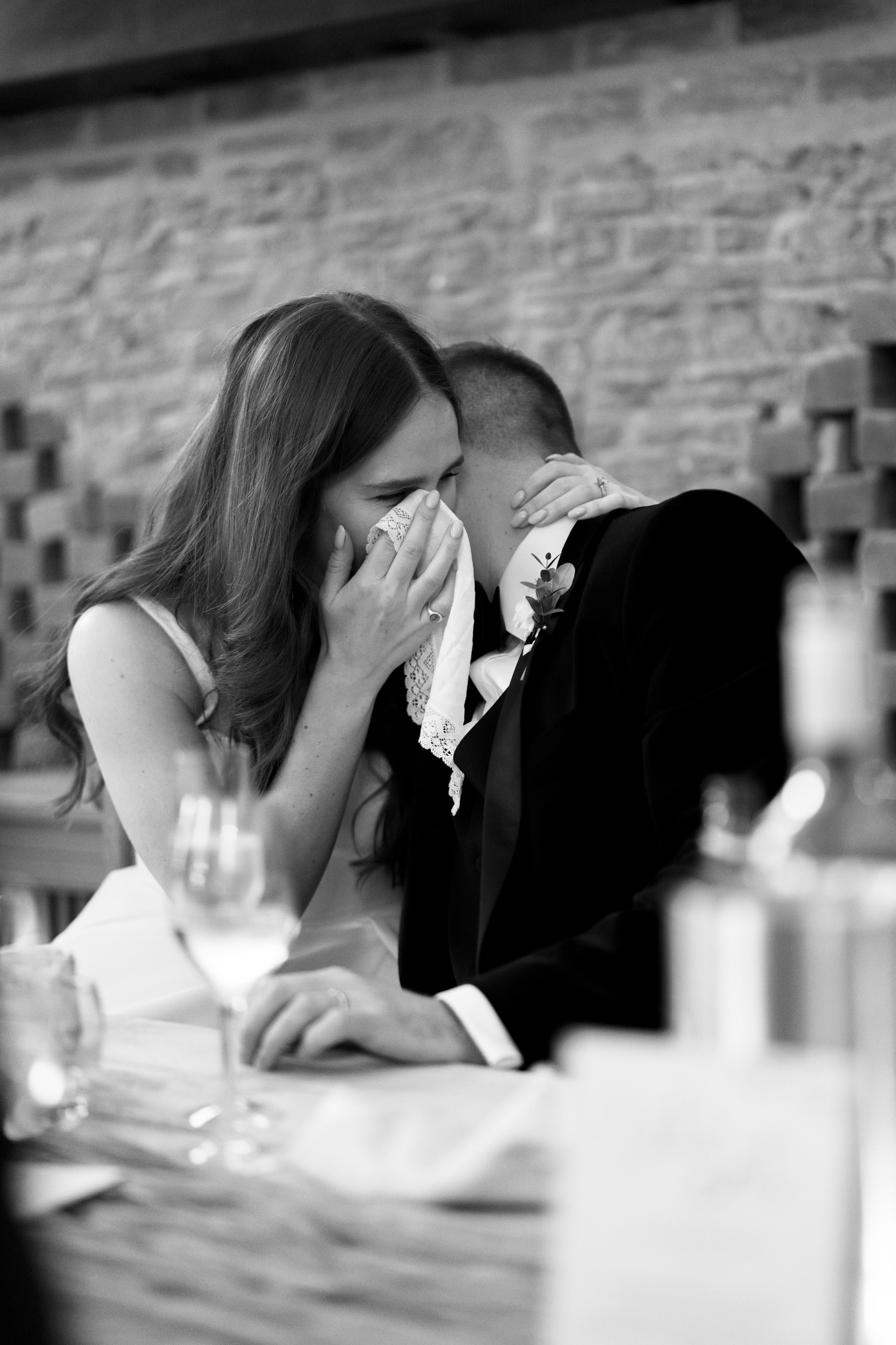 A bride and groom dressed in formal clothing sit together at a table. The bride seems emotional, holding a handkerchief to her face. There are wine glasses and bottles on the table.