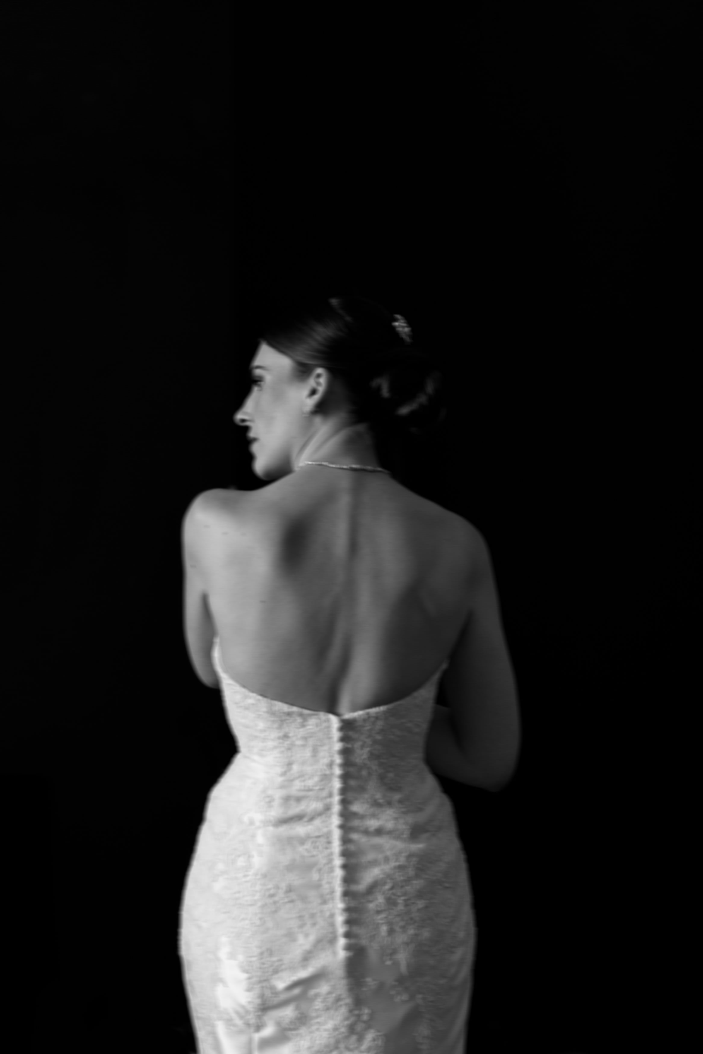 Someone wearing a backless, strapless dress is standing sideways in front of a dark background.