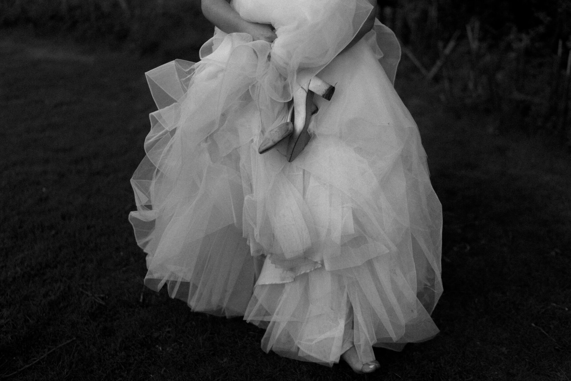 Someone is holding another person who is wearing a ruffled wedding dress and carrying high-heeled shoes, while standing on the grass.