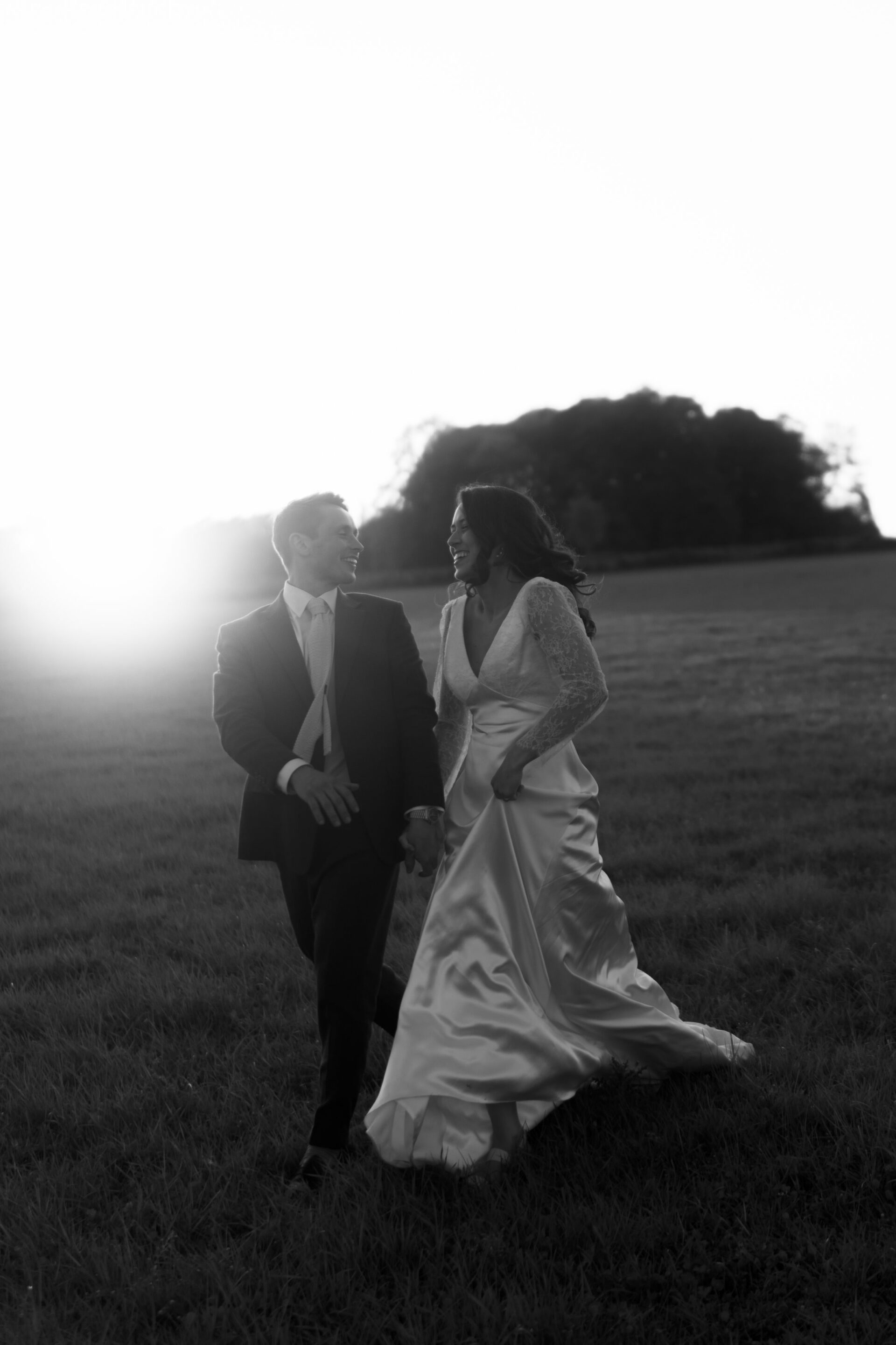 A couple dressed in their wedding outfits walks through a grassy field as the sun sets.