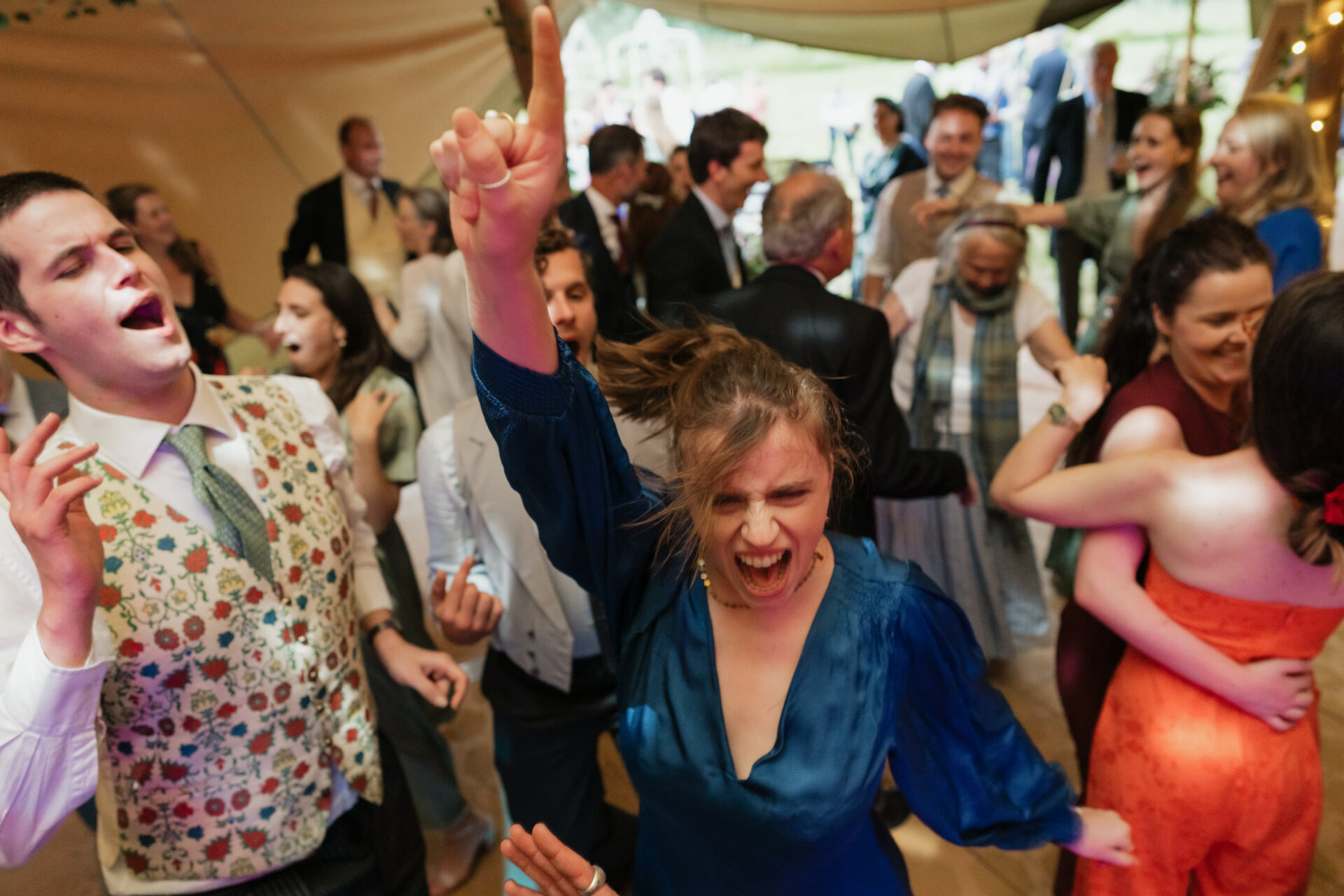 A group of people dancing energetically at a fun event, with a woman in a blue dress at the front raising her arm.