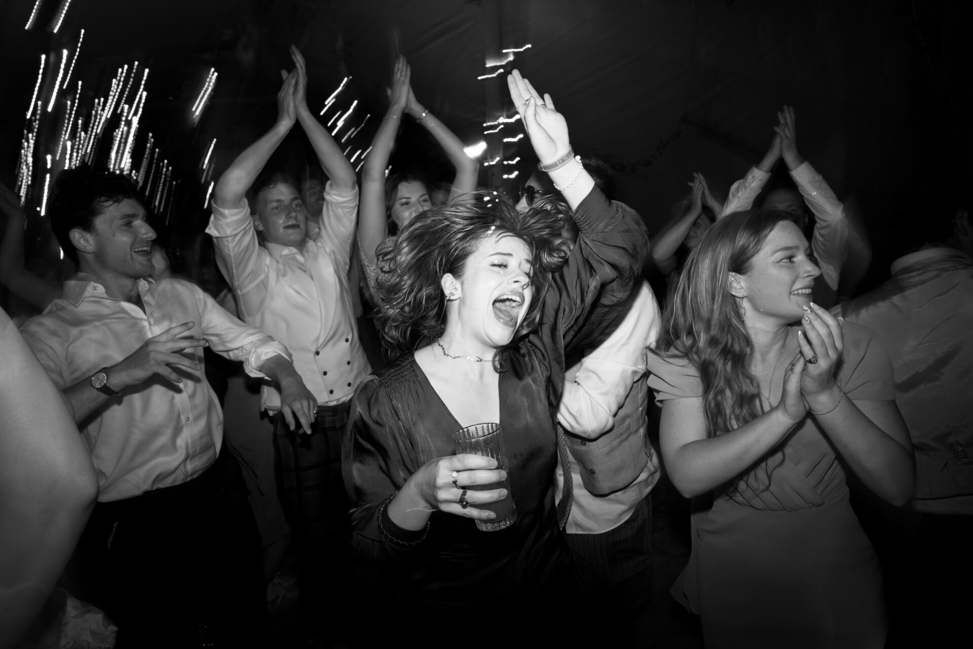 A bunch of people are excitedly dancing and clapping at a fun party, and there's one woman in the middle holding a drink.