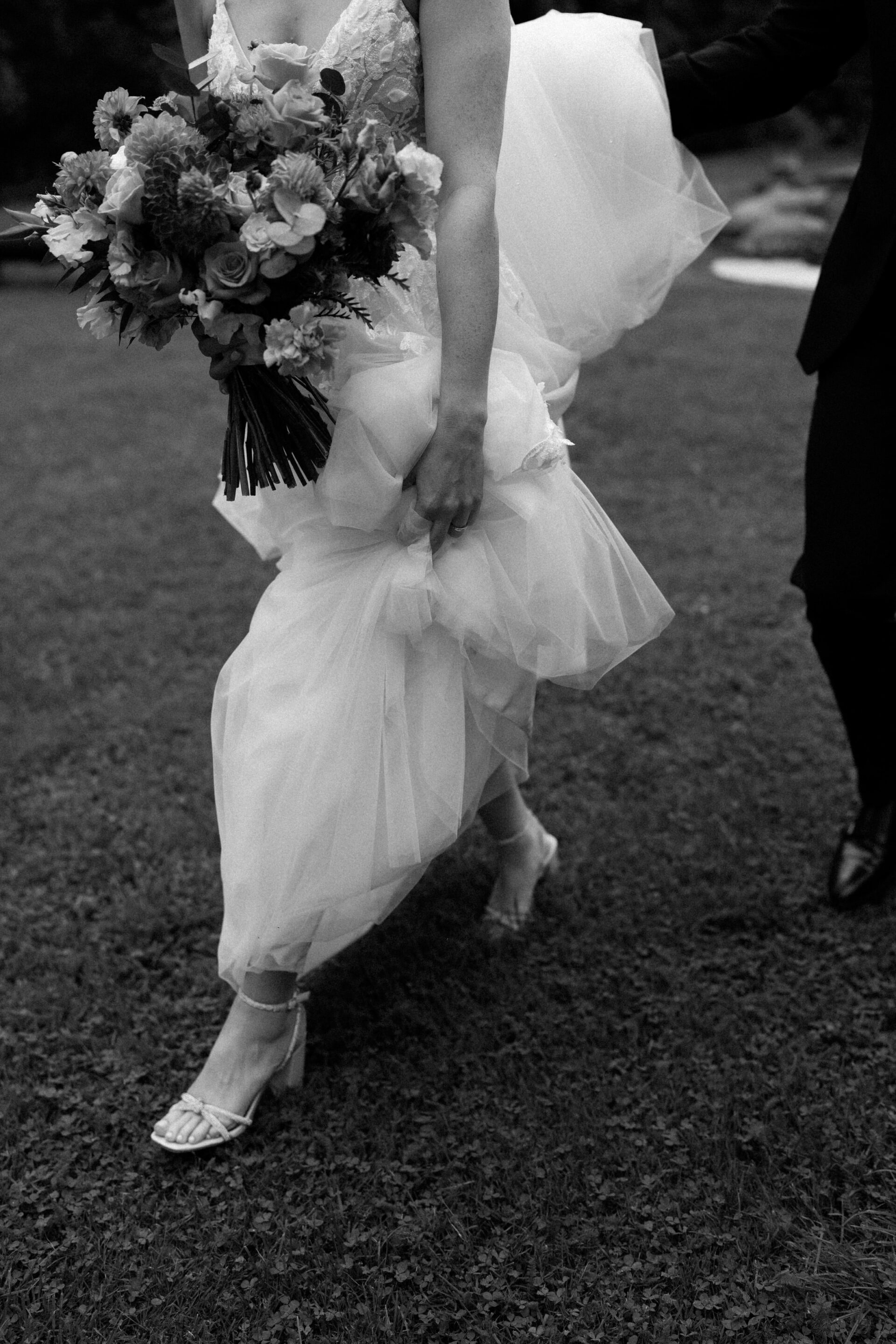 A bride is holding her bouquet and lifting her wedding dress as she walks on the grass.