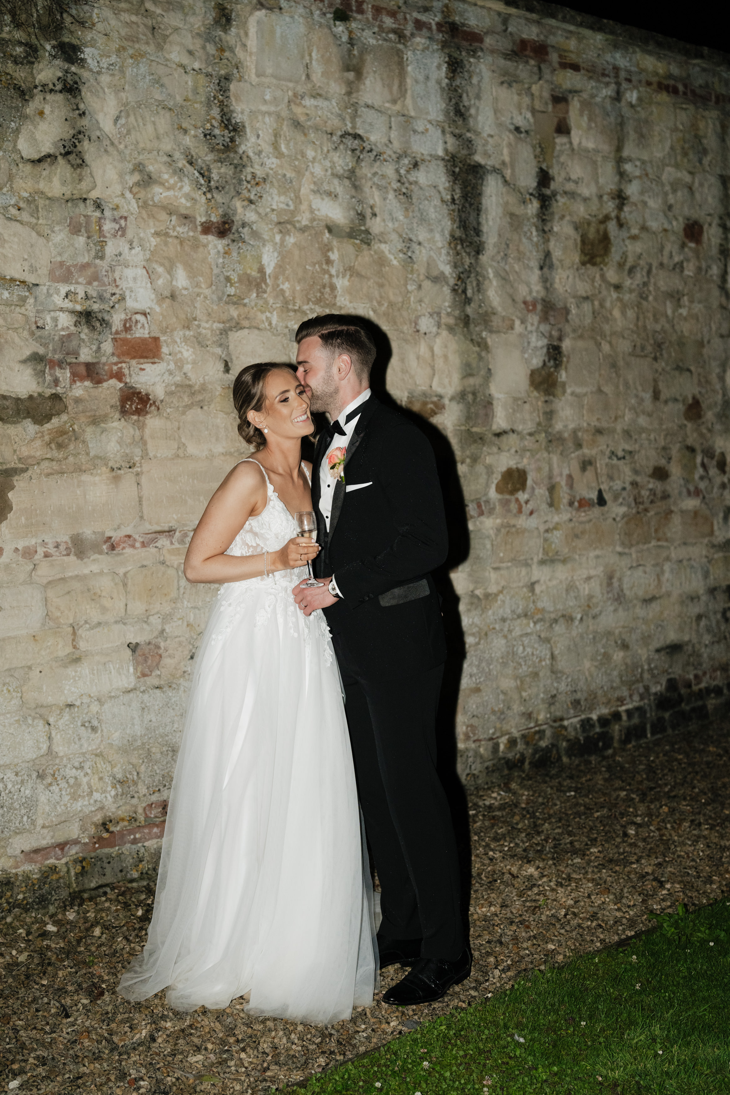 A couple dressed up for a special occasion stands in front of a stone wall at night. The woman is in a white dress, and the man is wearing a black tuxedo. They seem to be enjoying some time together.