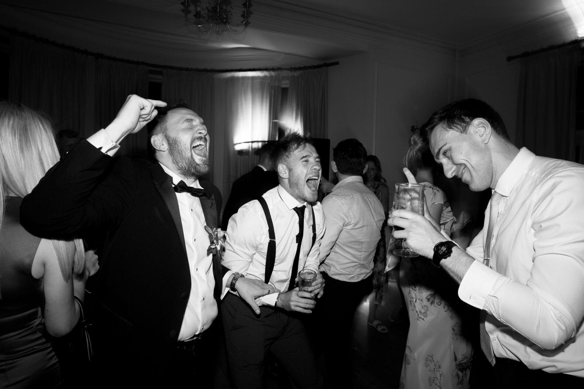 Three men in suits are laughing and using lots of hand gestures while having drinks at a party.