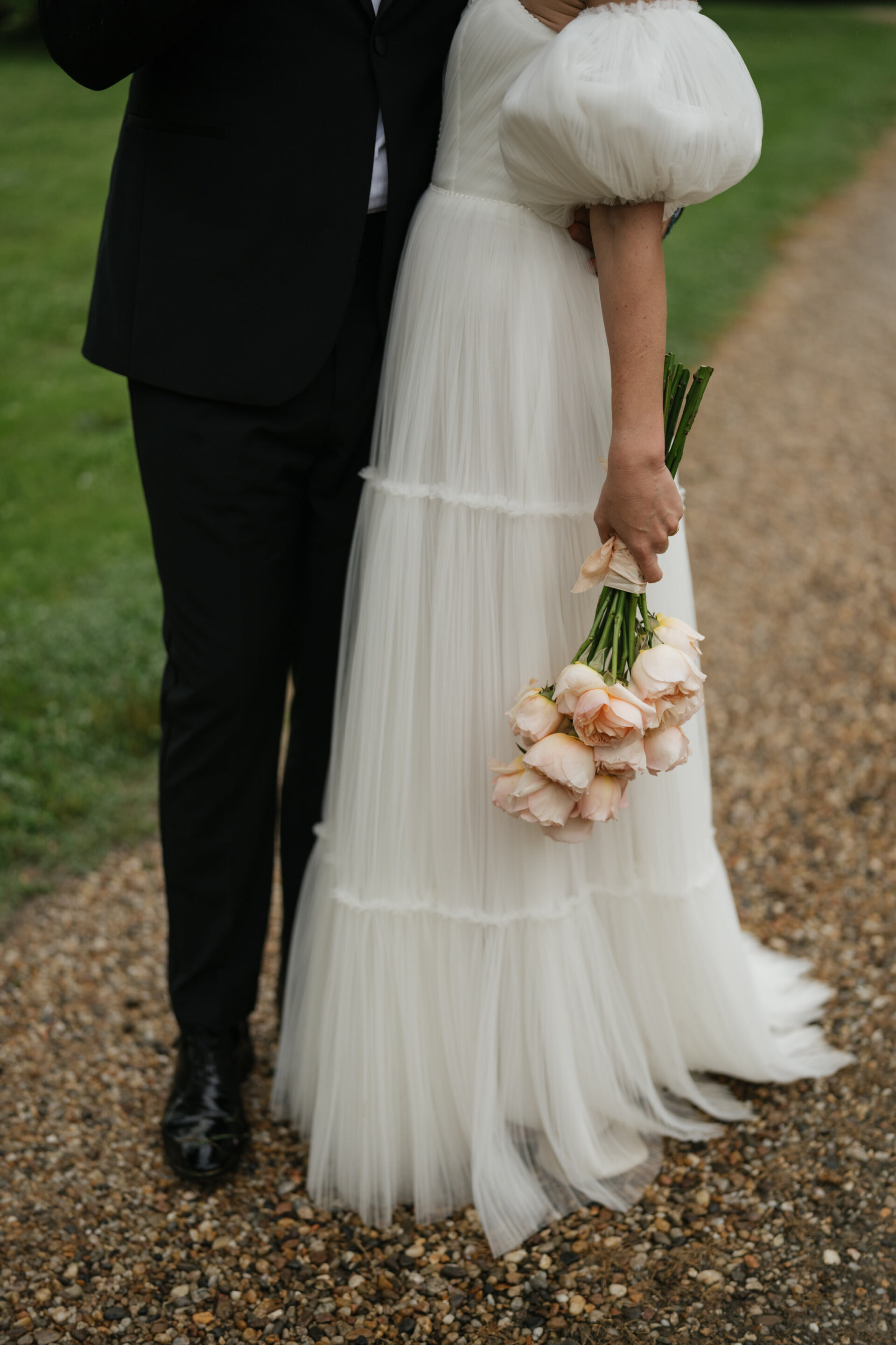 A couple is standing on a gravel path. The woman is wearing a white dress and holding a bunch of pink roses, while the man is dressed in a black suit.