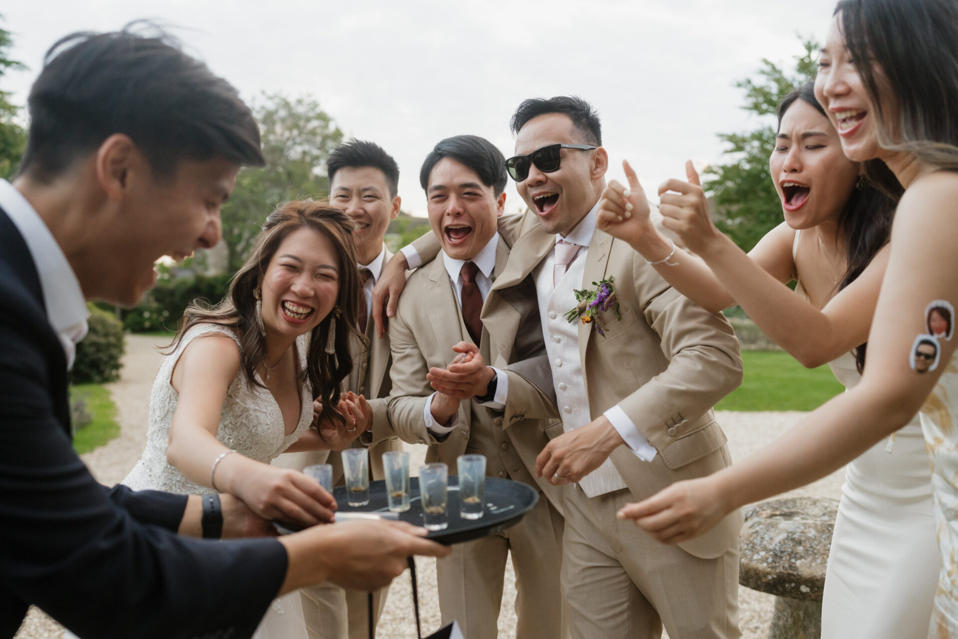 A group of nicely dressed people is outside, laughing and having fun together around someone who is holding a tray with shot glasses.