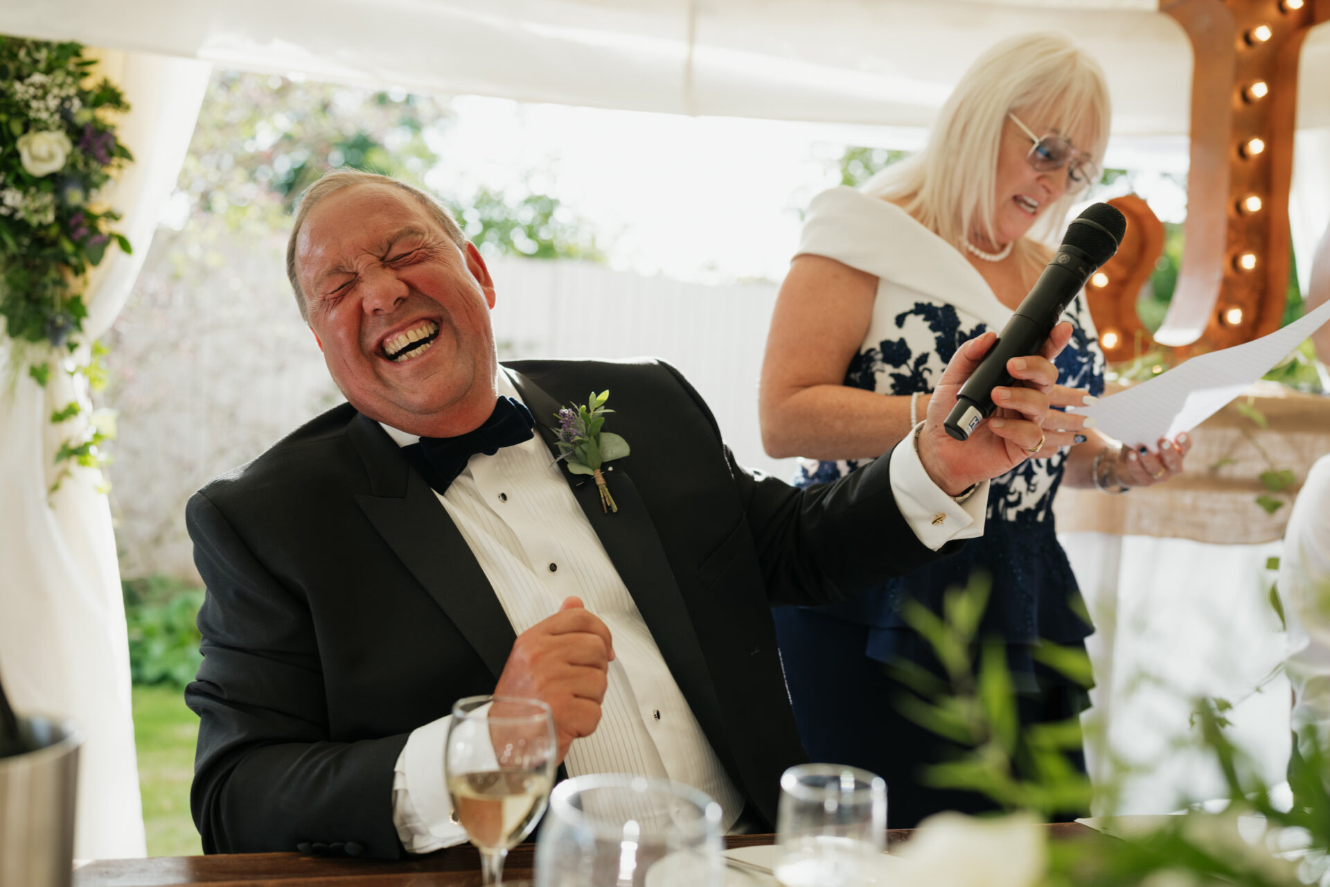 A man in a fancy suit is laughing loudly while holding a microphone. A woman next to him is reading from a piece of paper. It looks like they are at a formal event with nice decorations.