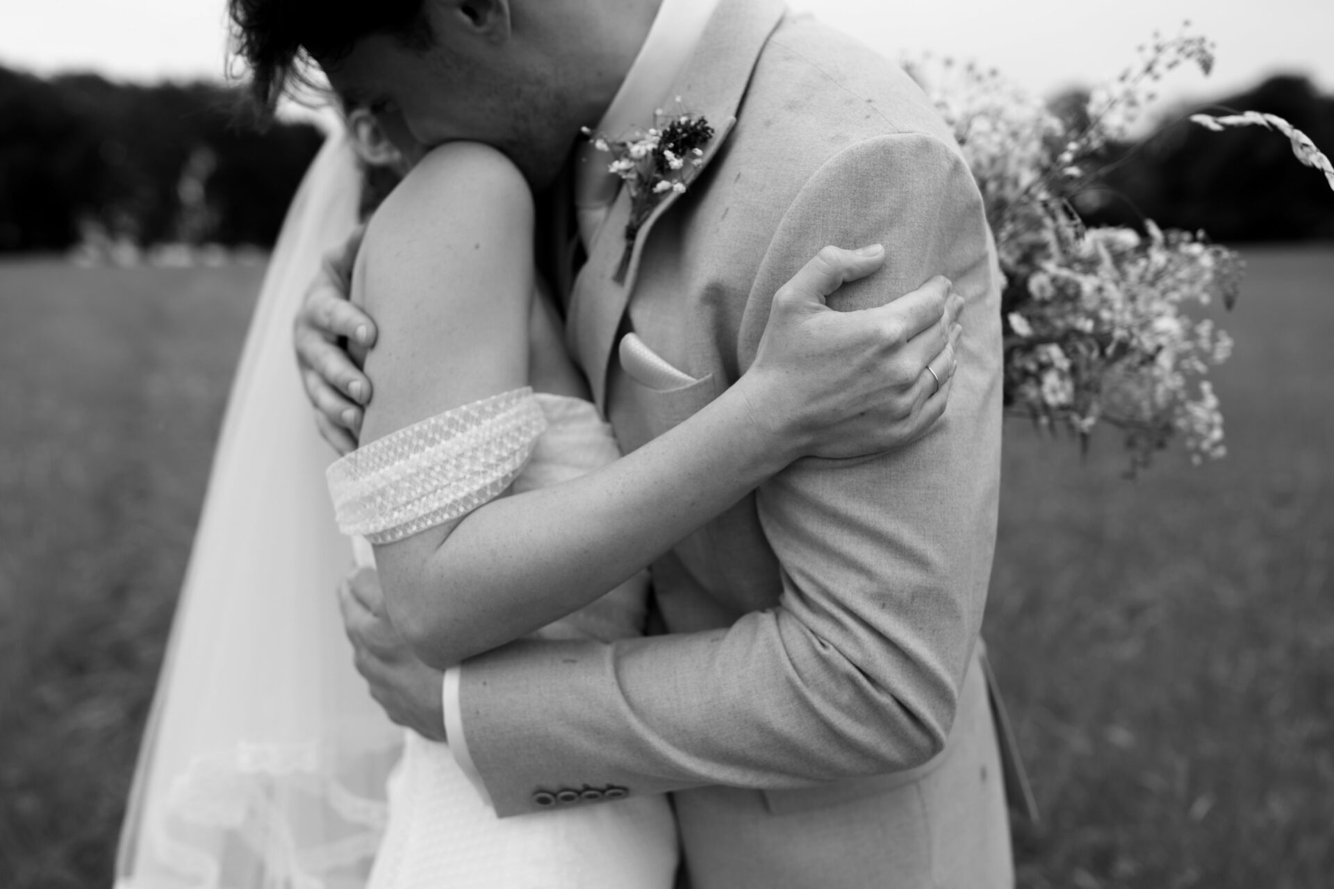 A bride and groom are hugging each other in a field. The groom is dressed in a suit, and the bride is holding a bunch of flowers.