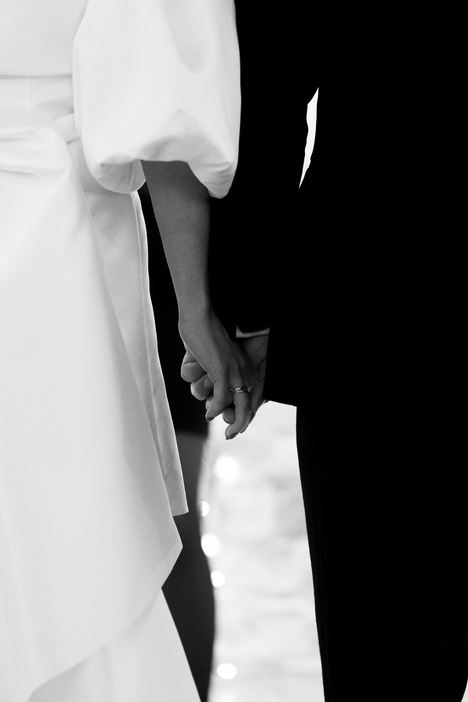 A bride and groom are holding hands and facing away from the camera in a black and white picture. The bride is wearing a dress with puffy sleeves, while the groom is dressed in a dark suit.