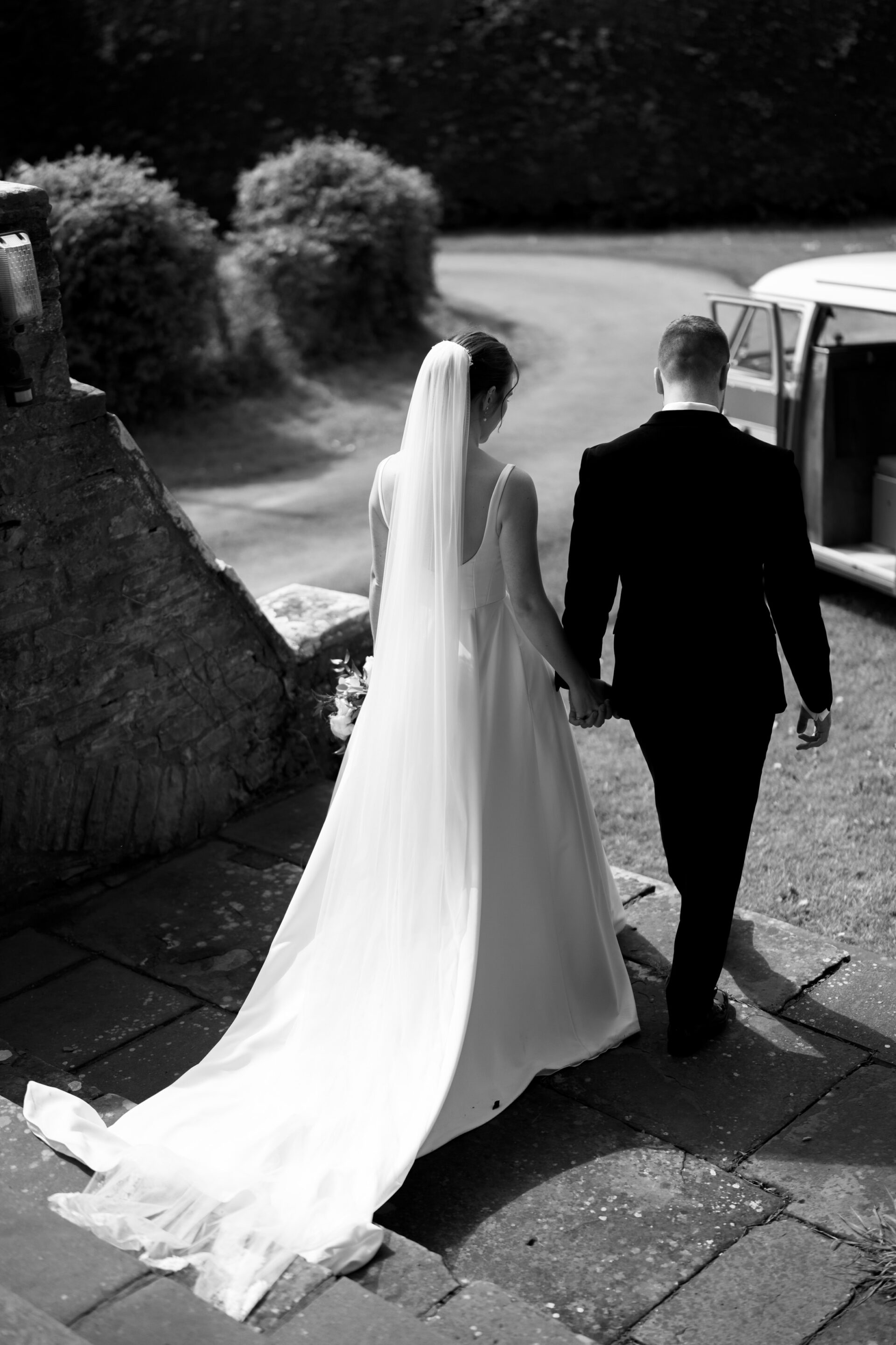 A bride and groom are walking together outside, holding hands. The bride is wearing a long dress and veil, and the groom is dressed in a dark suit.