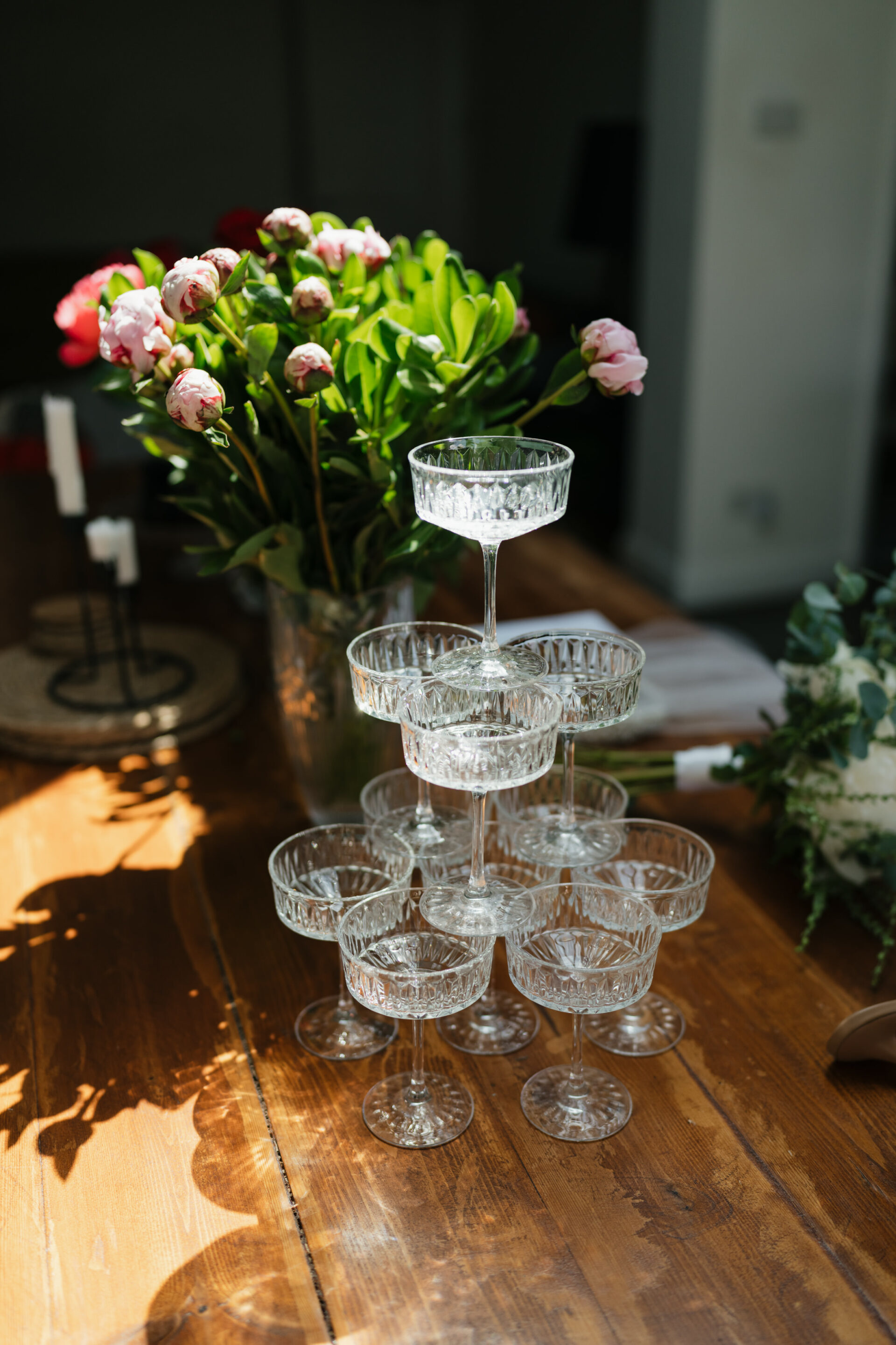A stack of empty crystal champagne glasses on a wooden table, with a bunch of pink flowers and greenery in the background.