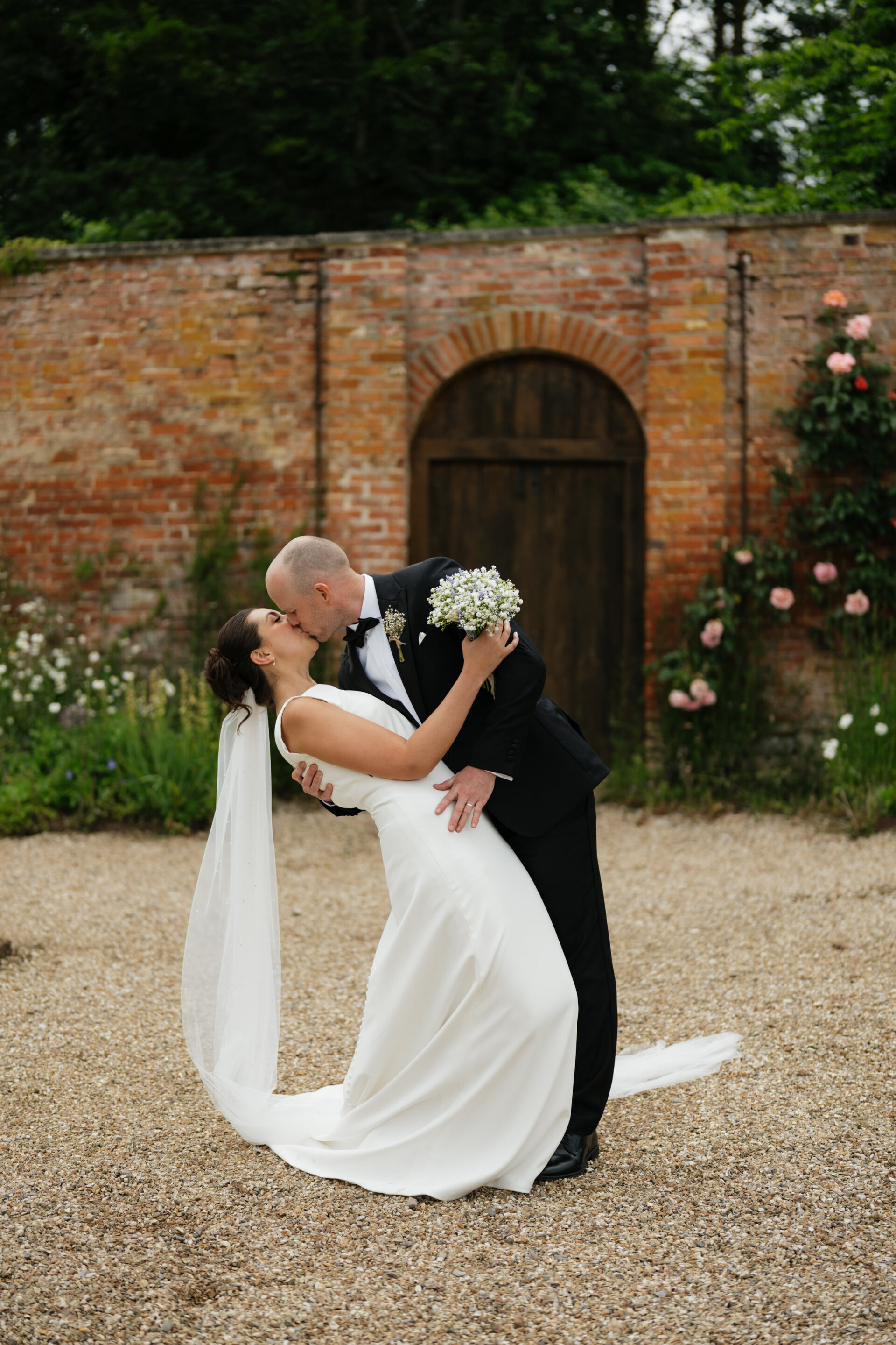 A bride and groom share a kiss in front of an old brick wall decorated with roses. The groom is dressed in a black suit, while the bride wears a white dress with a long veil and holds a bouquet of flowers.
